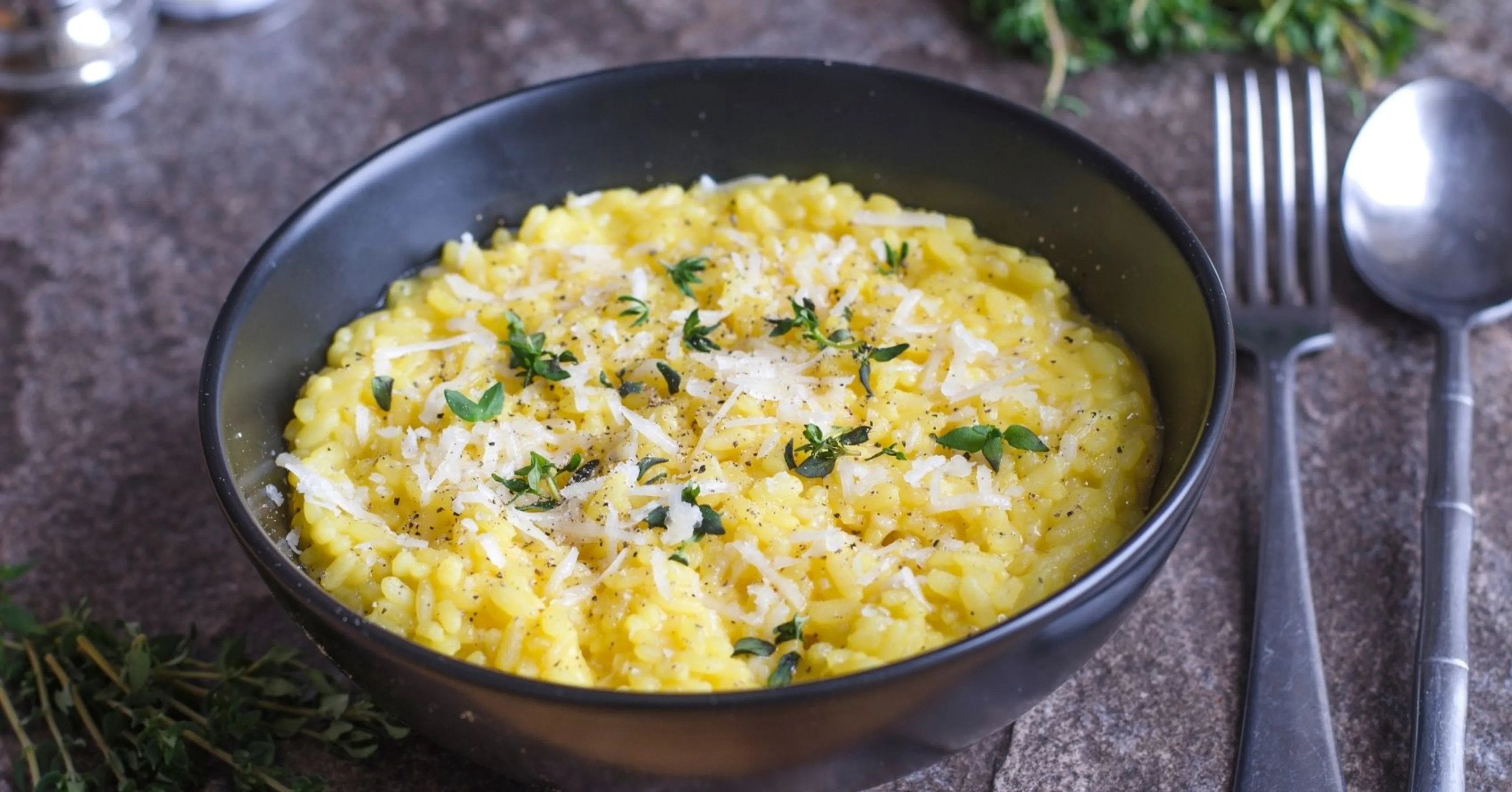 Krämig saffransrisotto toppad med riven parmesan och färsk timjan i en svart skål. Bredvid ligger en gaffel och en sked på en stenmönstrad yta.