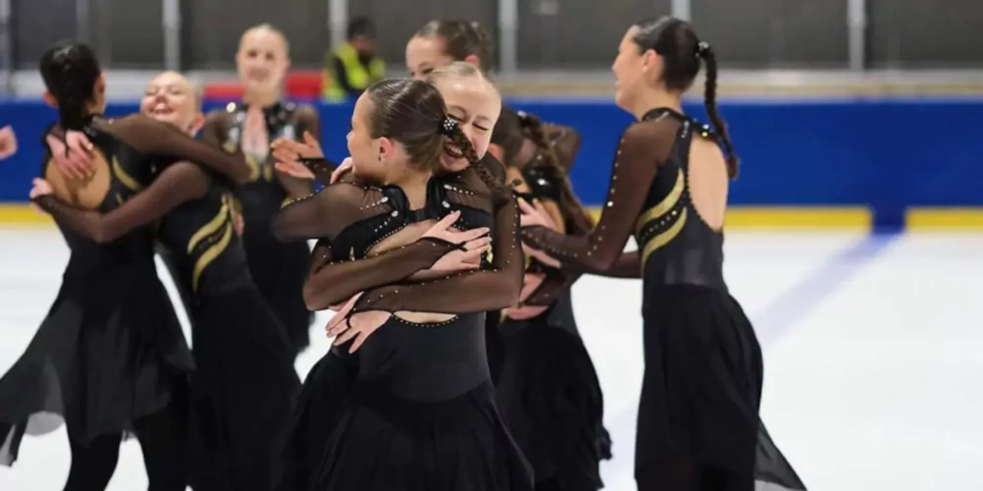 Ett team av unga skridskotjejer i svarta, glittriga dräkter kramas på isen efter en uppvisning. Glädje och samhörighet syns tydligt i deras leenden och kroppsspråk.