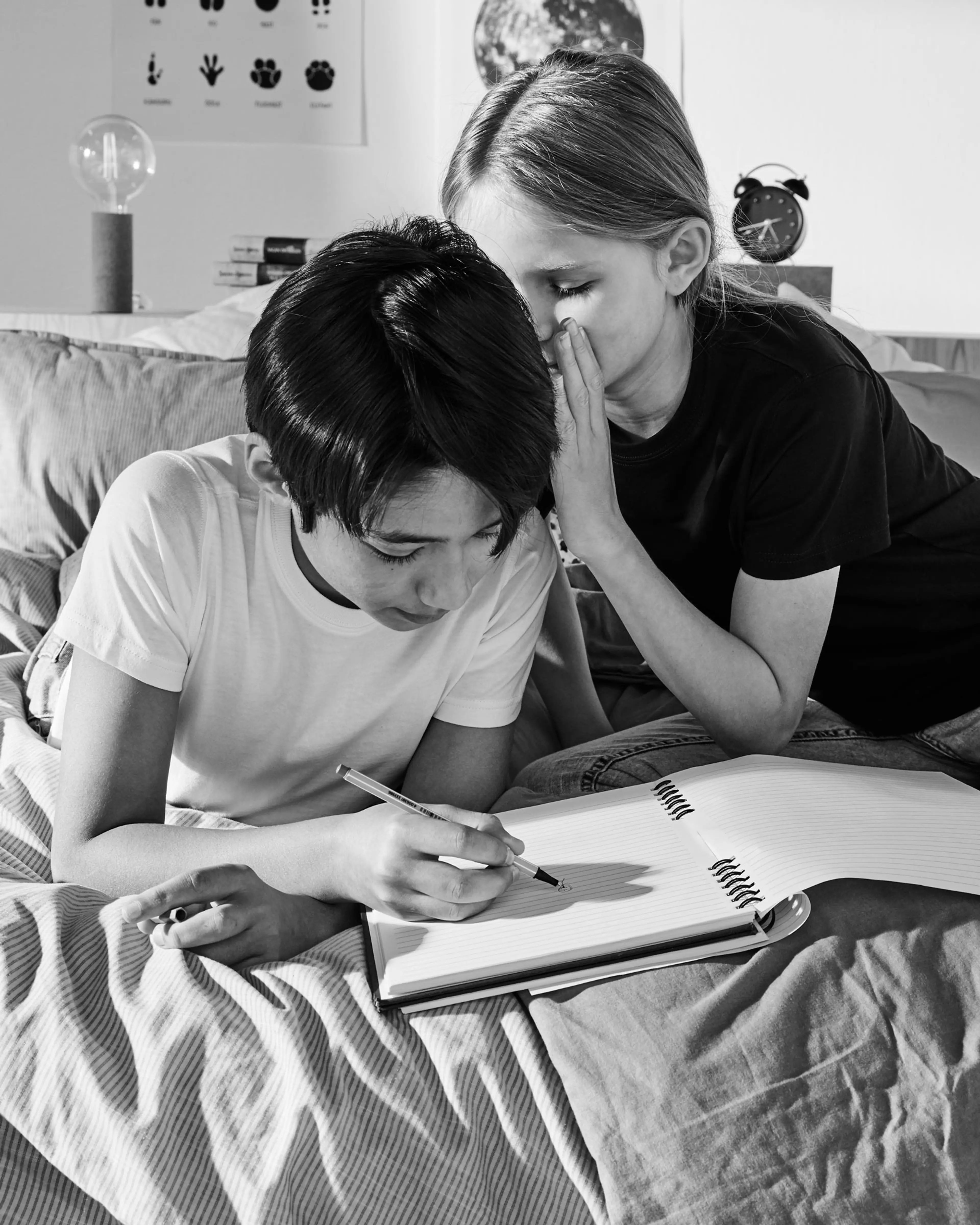 Två barn sitter i en säng där en viskar något i den andres öra medan den skriver i en spiralbunden anteckningsbok. De bär enkla t-shirtar i svart och vitt.