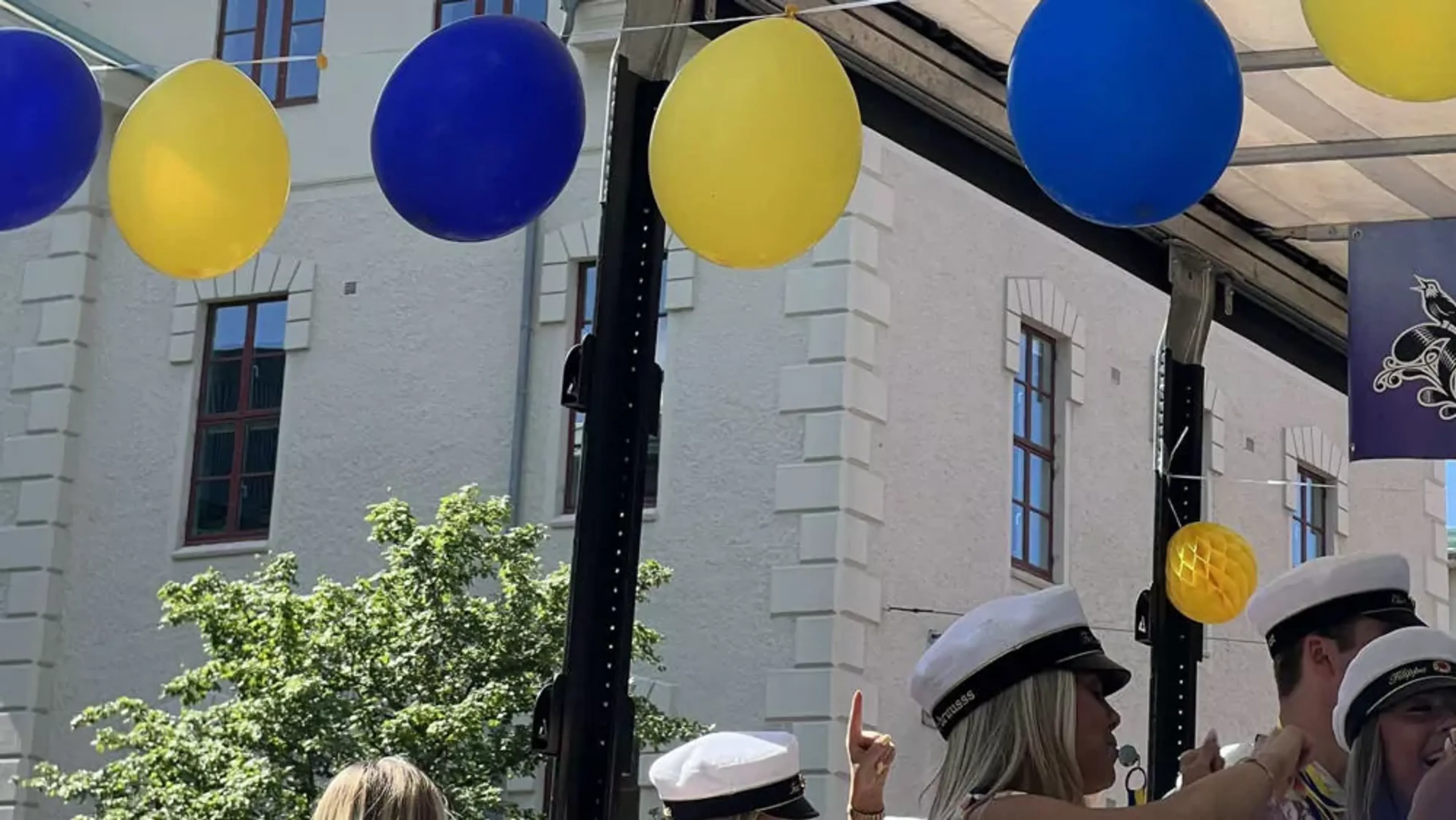 Studentfirande på ett flak med gula och blå ballonger, där flera personer bär traditionella studentmössor. Solen lyser och en byggnad syns i bakgrunden.