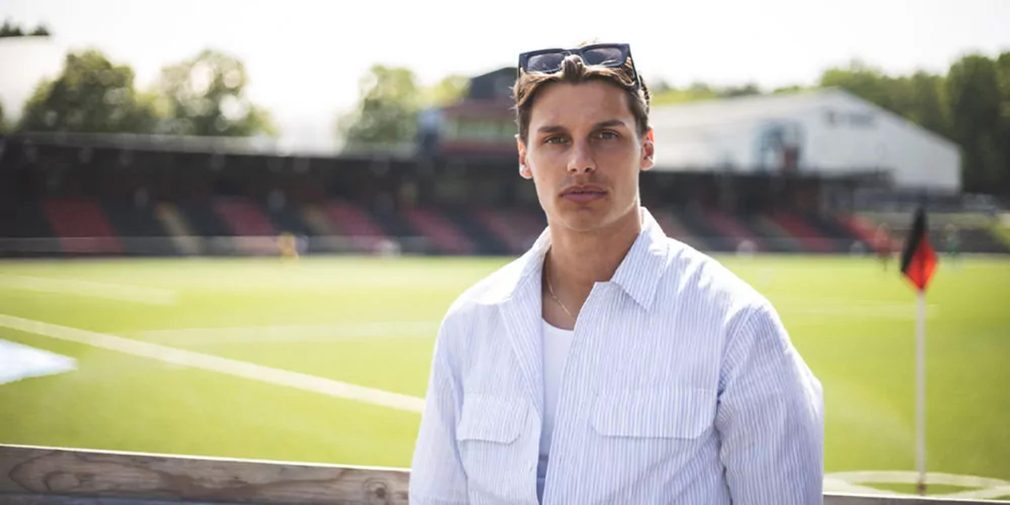 En man i ljusblå skjorta och solglasögon i håret står vid ett stängsel med en fotbollsplan i bakgrunden. Ljuset är mjukt och det är en solig dag.