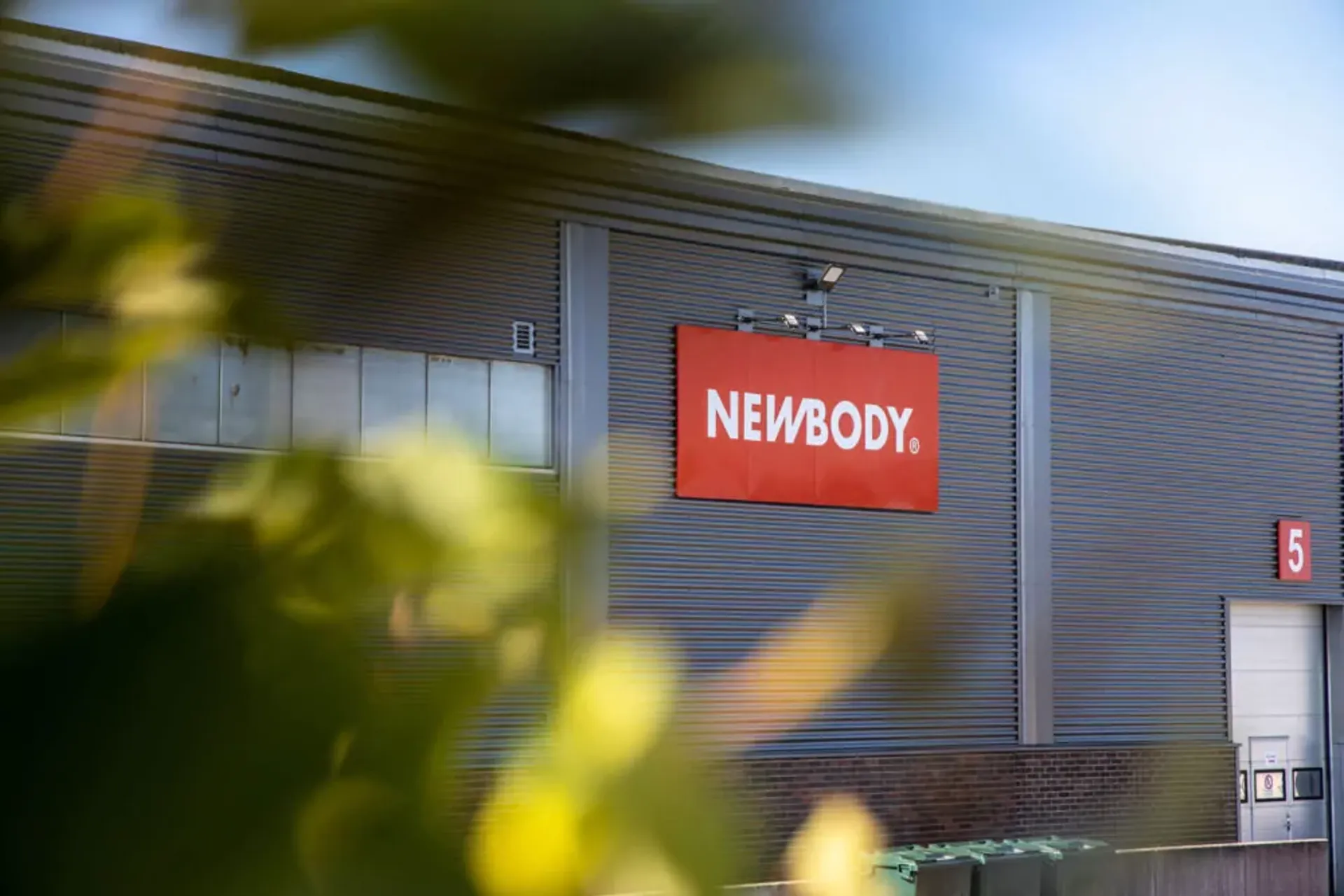 En industribyggnad i grått med en röd skylt på fasaden och numret 5 vid en vit lastport. Gröna löv i förgrunden skapar en mjuk kontrast mot byggnadens raka linjer.