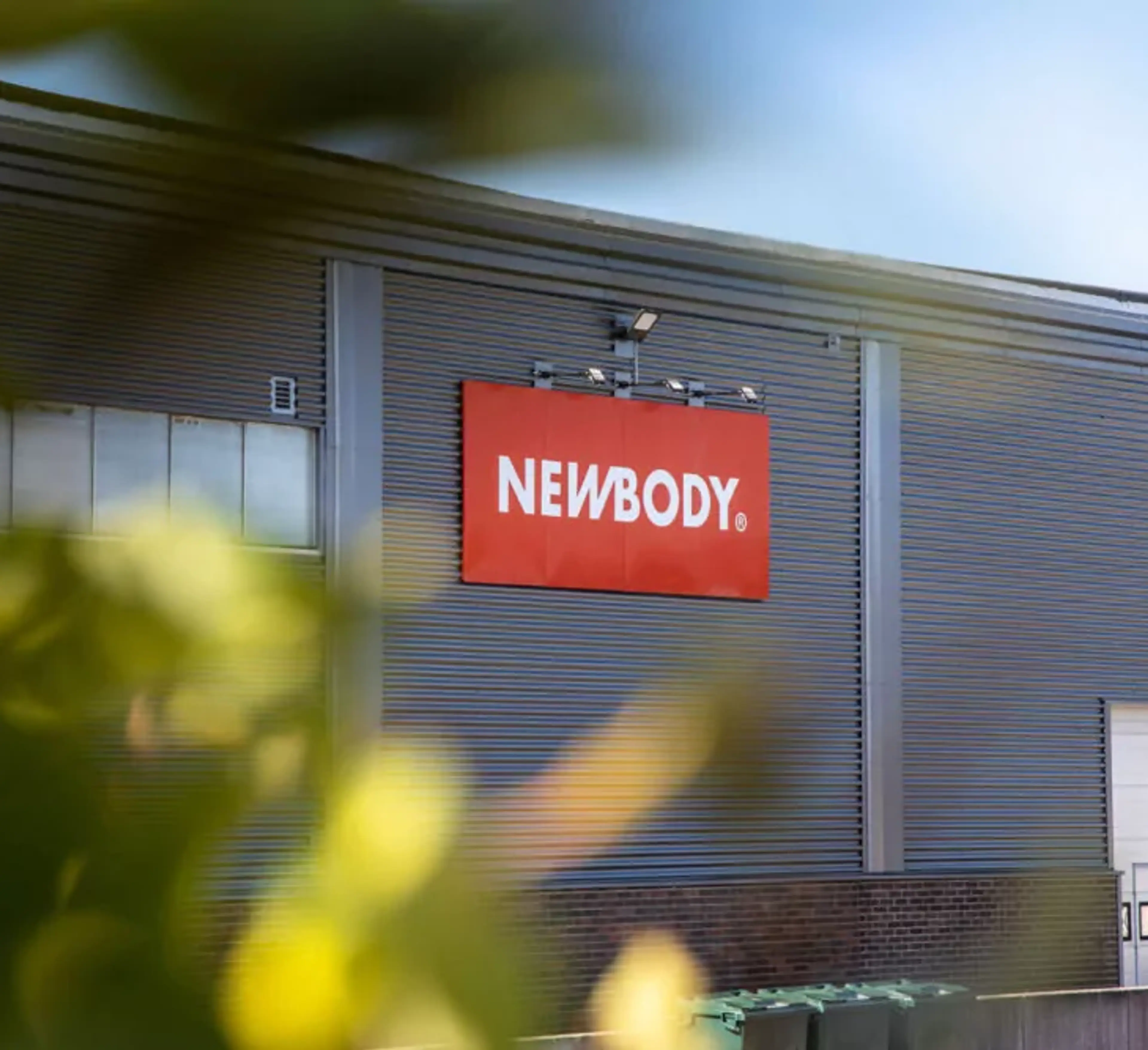 En industribyggnad i grått med en röd skylt på fasaden och numret 5 vid en vit lastport. Gröna löv i förgrunden skapar en mjuk kontrast mot byggnadens raka linjer.