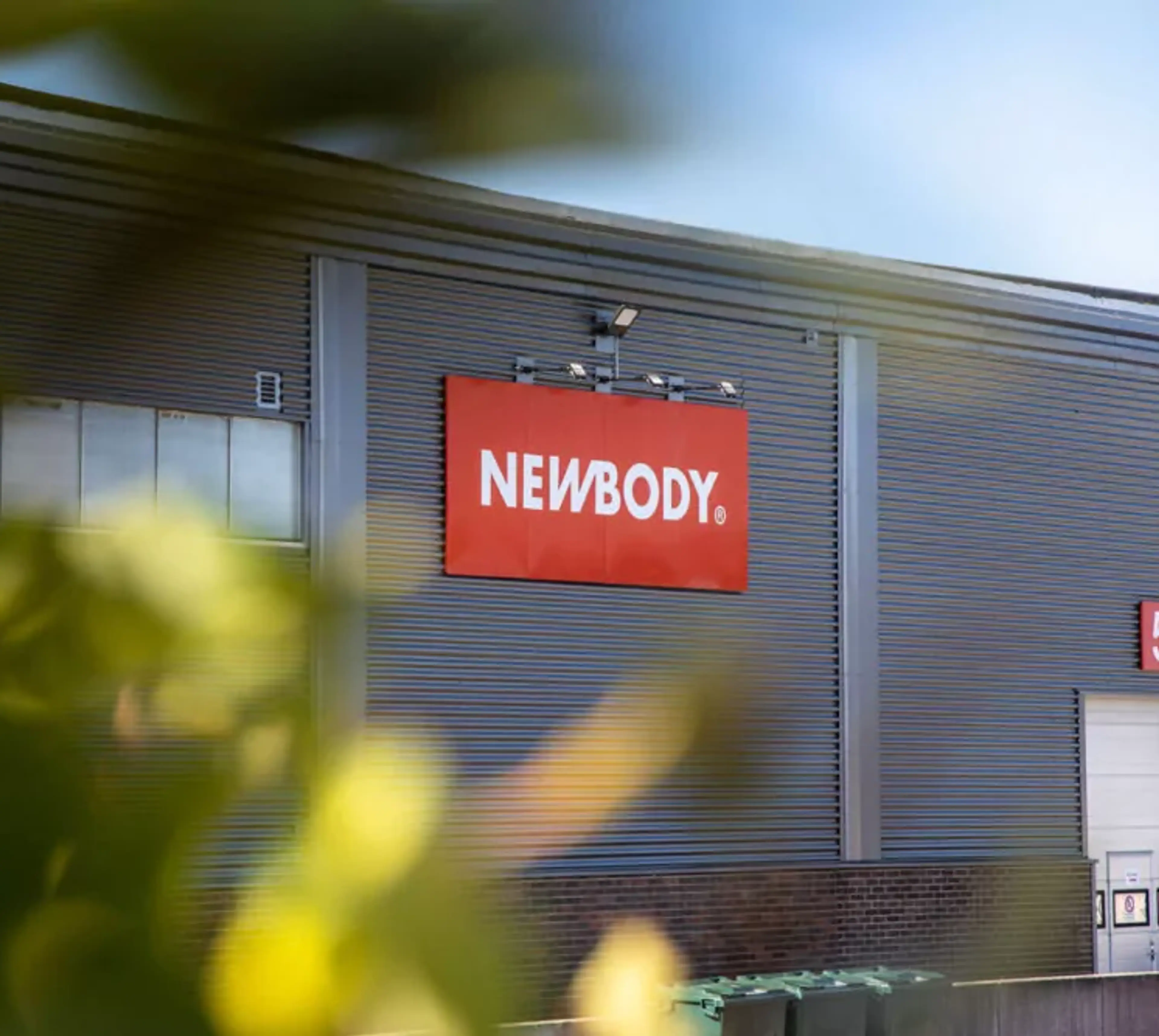 En industribyggnad i grått med en röd skylt på fasaden och numret 5 vid en vit lastport. Gröna löv i förgrunden skapar en mjuk kontrast mot byggnadens raka linjer.