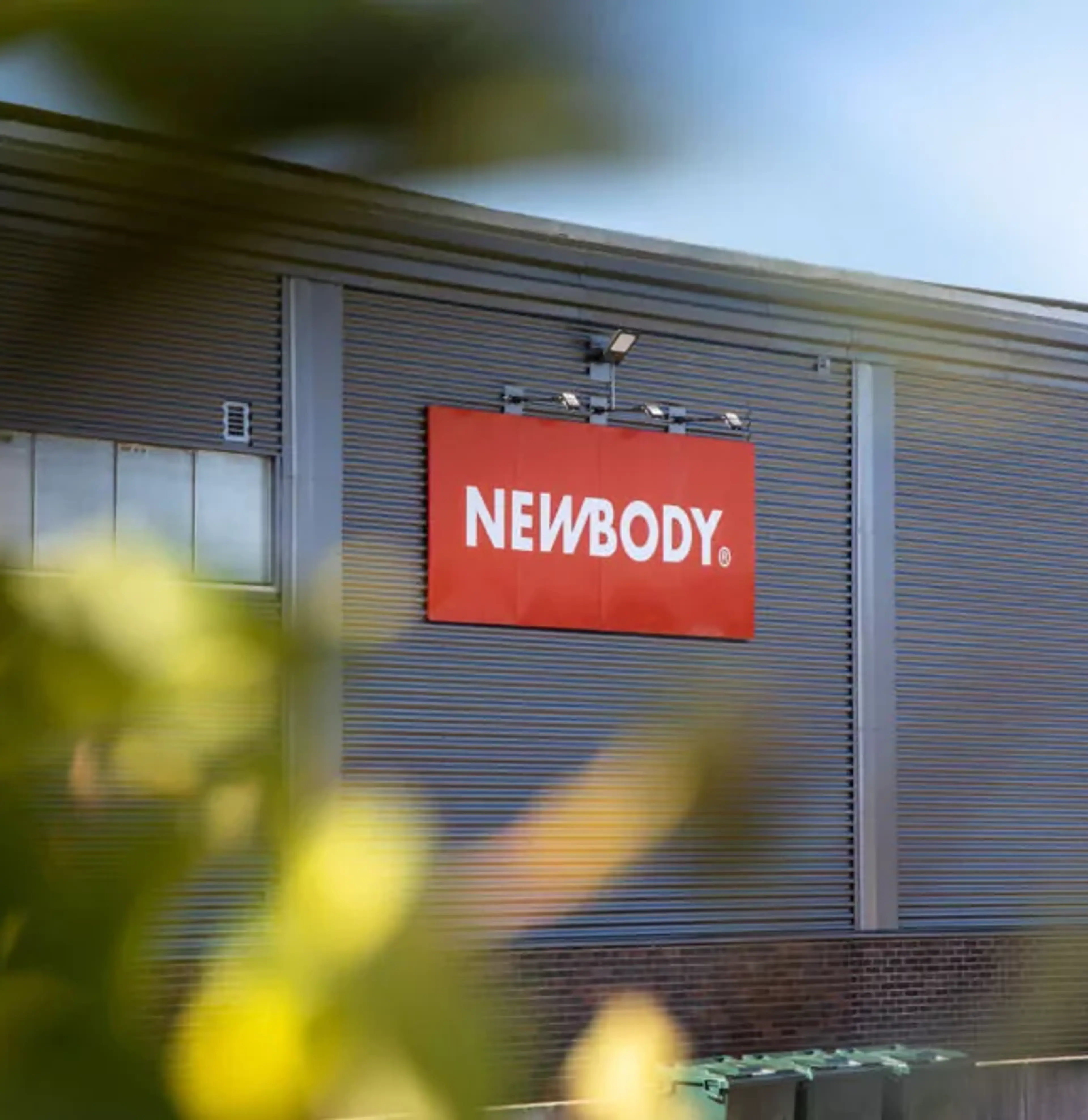 En industribyggnad i grått med en röd skylt på fasaden och numret 5 vid en vit lastport. Gröna löv i förgrunden skapar en mjuk kontrast mot byggnadens raka linjer.