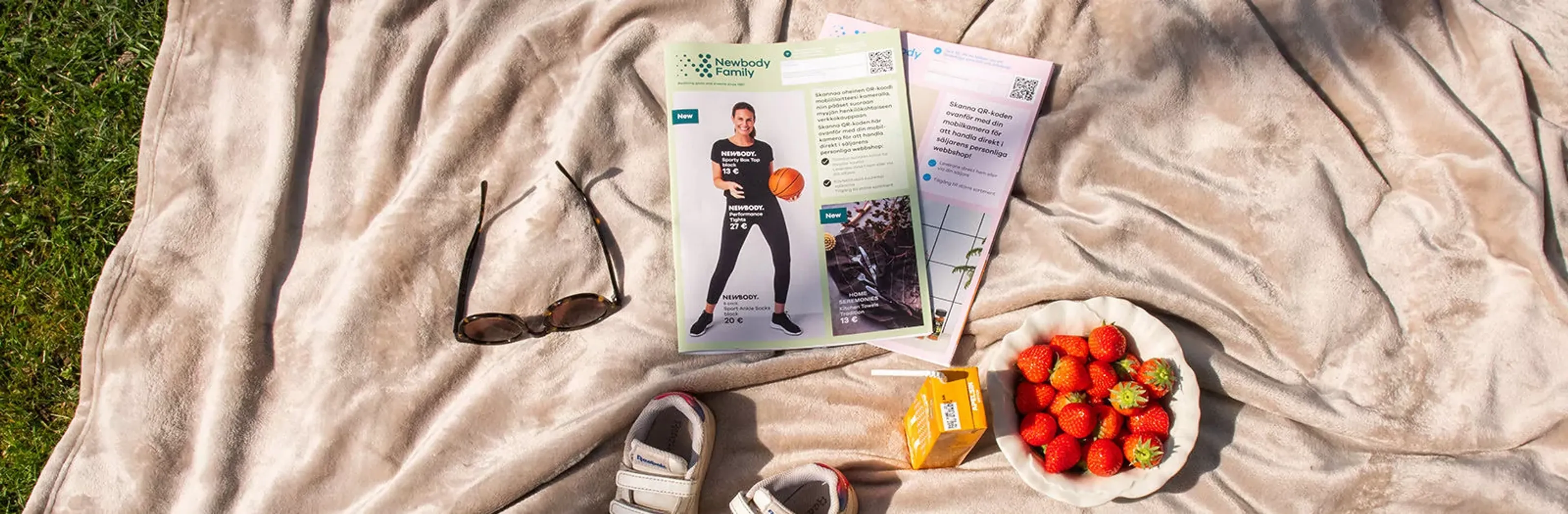 En mjuk, beige filt ligger utbredd på gräs med en barnkatalog, solglasögon, vita barnskor, en skål med jordgubbar och en juicekartong ovanpå.