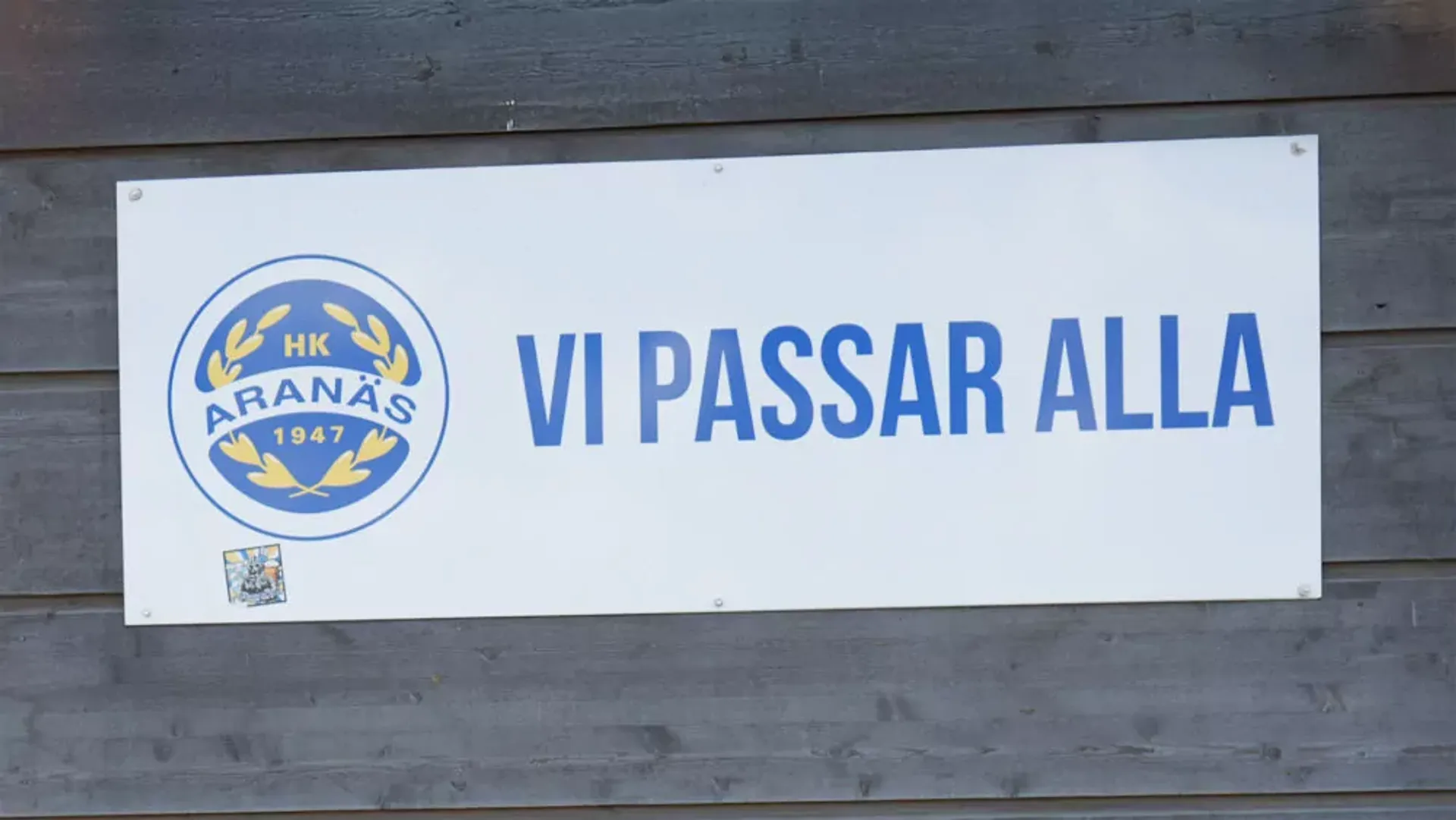En vit skylt med blå text där det står "Vi passar alla" och en blå och gul logga med texten "HK Aranäs 1947". Skylten är monterad på en grå trävägg.