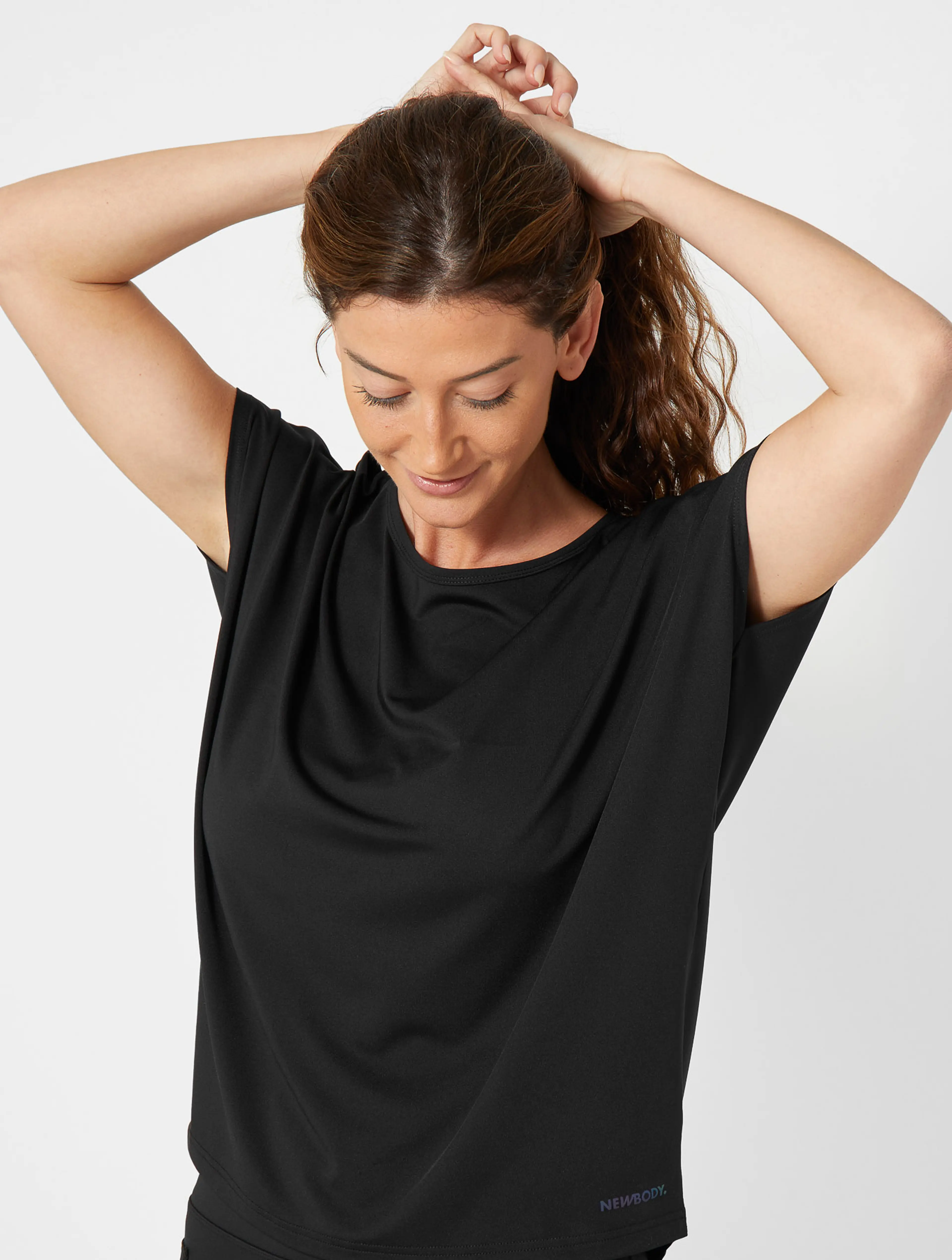 En svart, kortärmad sporttopp i mjukt och stretchigt material med rundad halsringning, bärs av en kvinna som just sätter upp sitt hår.