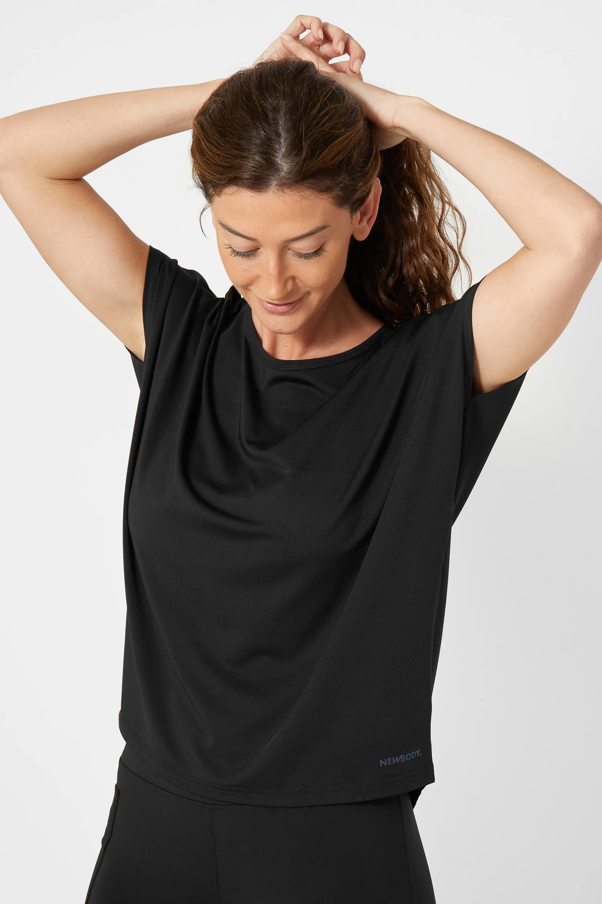 En svart, kortärmad sporttopp i mjukt och stretchigt material med rundad halsringning, bärs av en kvinna som just sätter upp sitt hår.