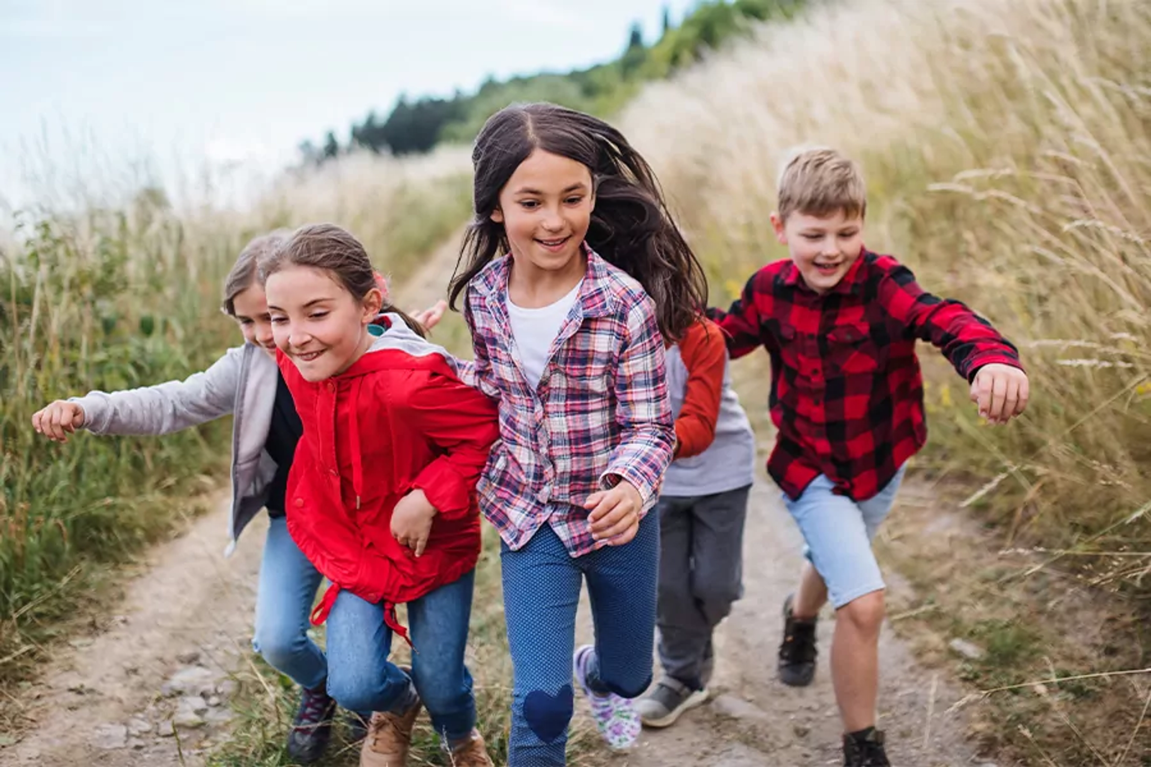 En grupp barn i färgglada kläder springer längs en grusväg kantad av högt gräs. Deras glada miner och rörelse fångar en fri och energisk stund utomhus.
