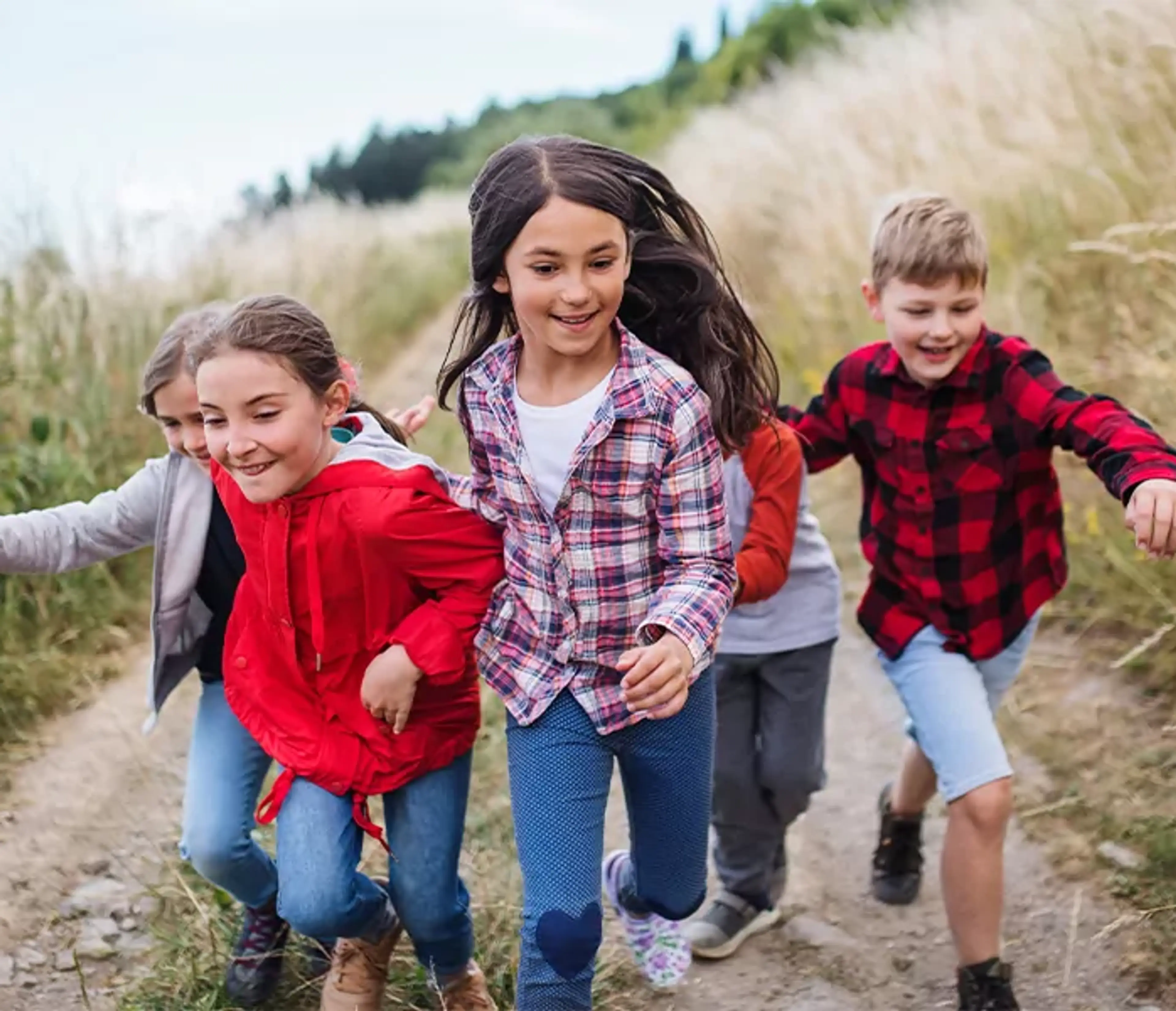 En grupp barn i färgglada kläder springer längs en grusväg kantad av högt gräs. Deras glada miner och rörelse fångar en fri och energisk stund utomhus.