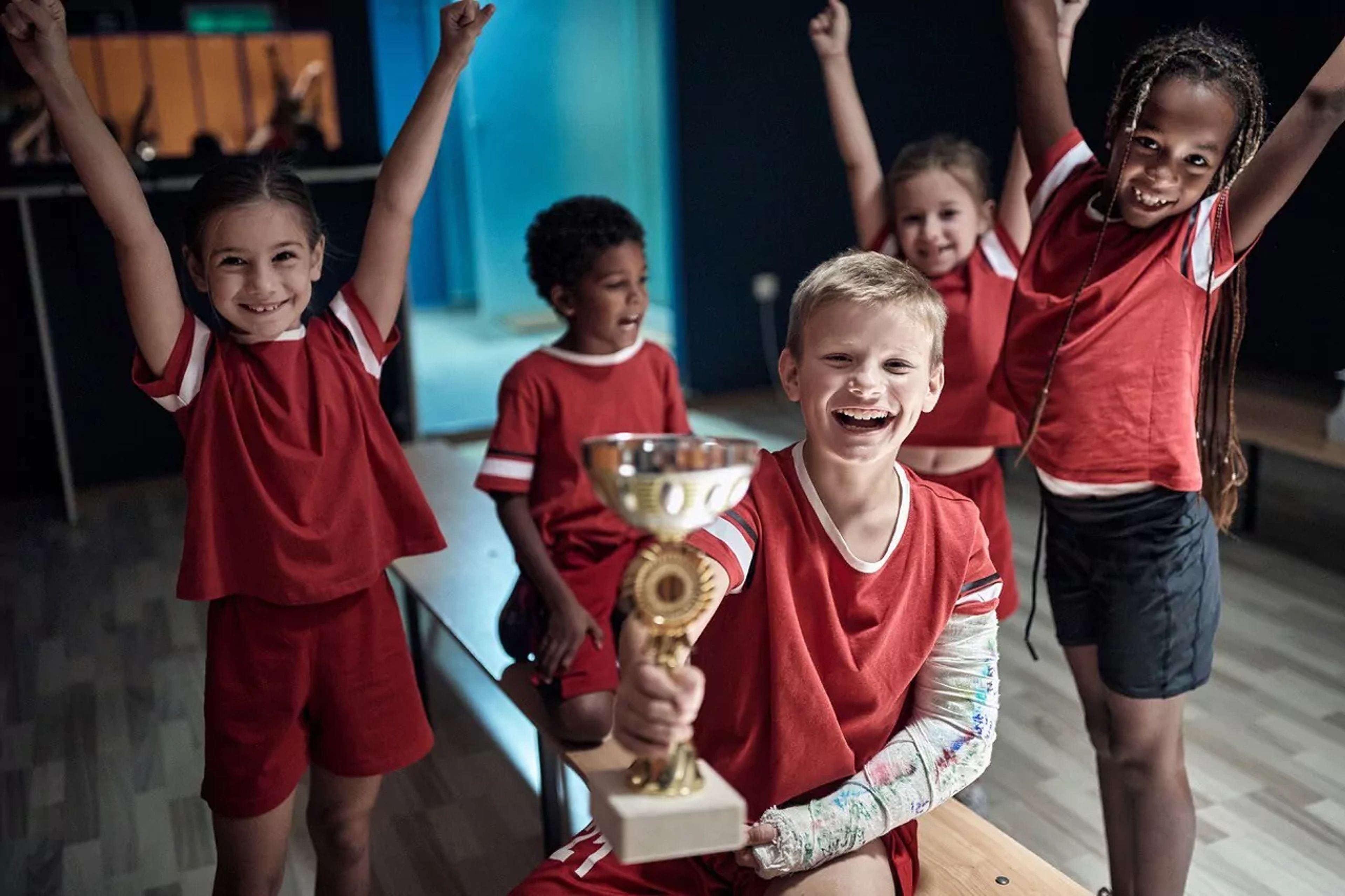 Fem barn i röda idrottskläder firar tillsammans, där en pojke med gipsad arm håller upp en pokal. Några av barnen sitter på en bänk medan andra står med armarna i luften.