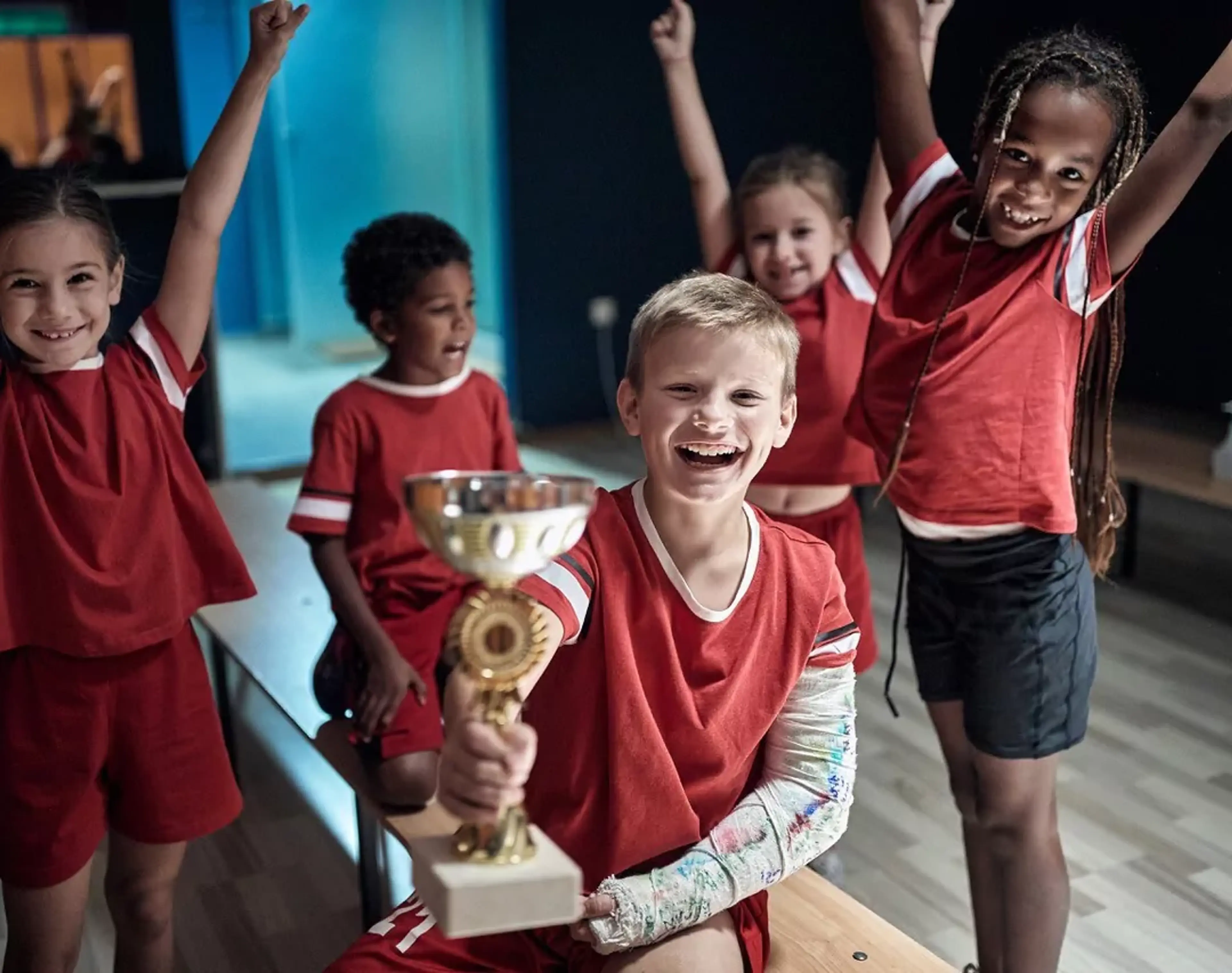 Fem barn i röda idrottskläder firar tillsammans, där en pojke med gipsad arm håller upp en pokal. Några av barnen sitter på en bänk medan andra står med armarna i luften.