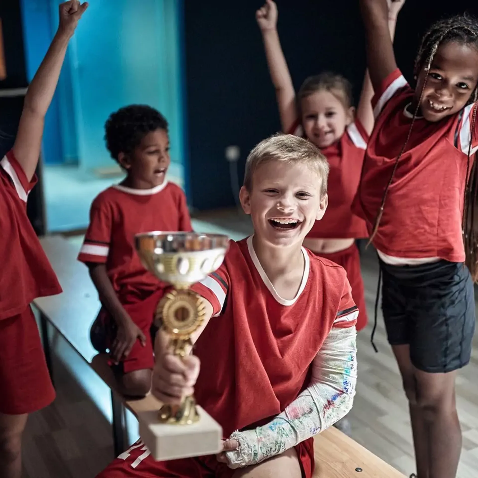 Fem barn i röda idrottskläder firar tillsammans, där en pojke med gipsad arm håller upp en pokal. Några av barnen sitter på en bänk medan andra står med armarna i luften.