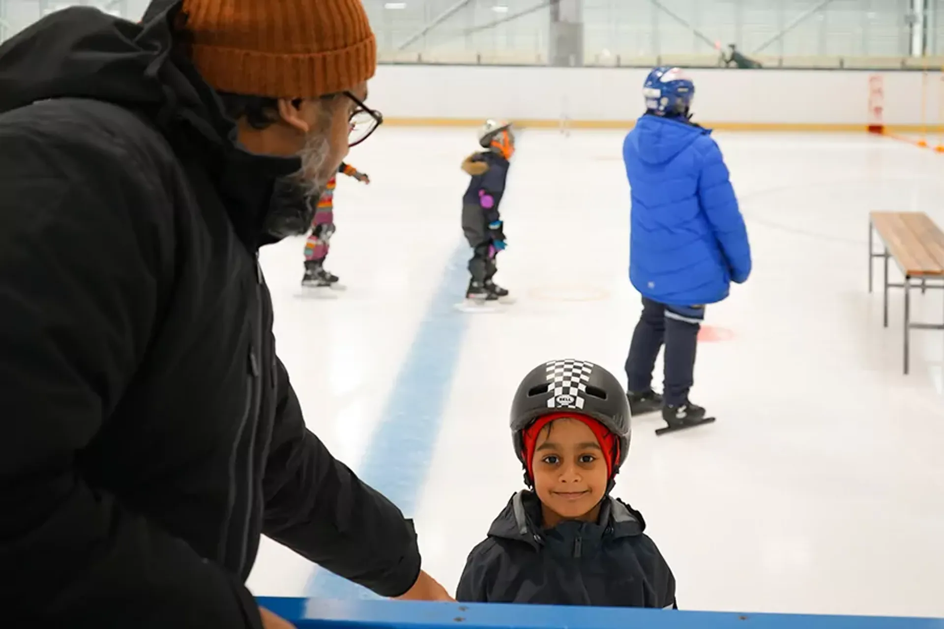Ett barn med hjälm och varm jacka står på isen och tittar mot kameran medan en vuxen i mössa och glasögon håller stöd vid sargen. Andra barn i vinterkläder åker skridskor i bakgrunden.