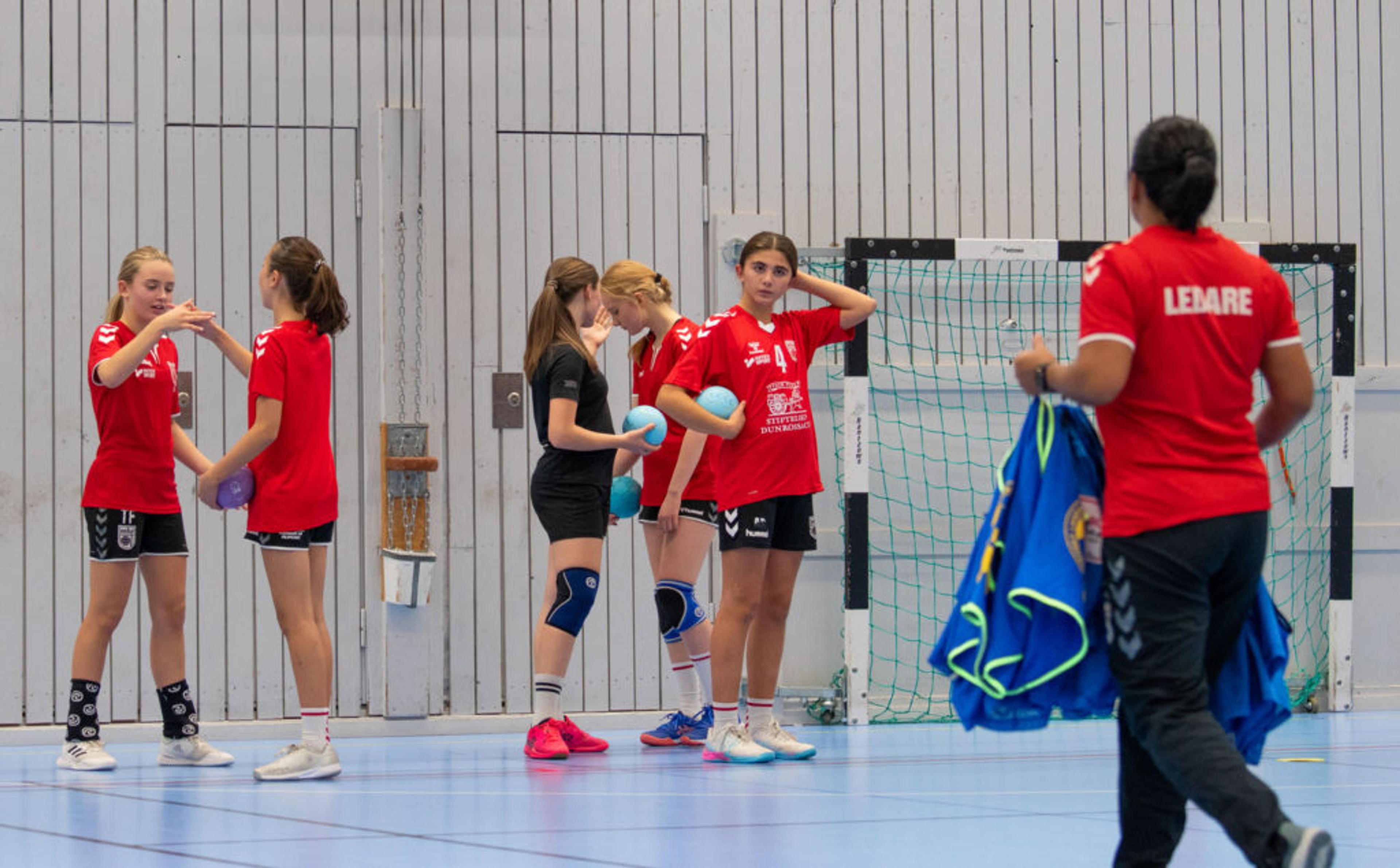 En grupp ungdomar i sportkläder samlas i en gymnastikhall med en basketkorg och innebandymål i bakgrunden. En ledare bär en röd tröja där det står "LEDARE" och håller i blå västar.