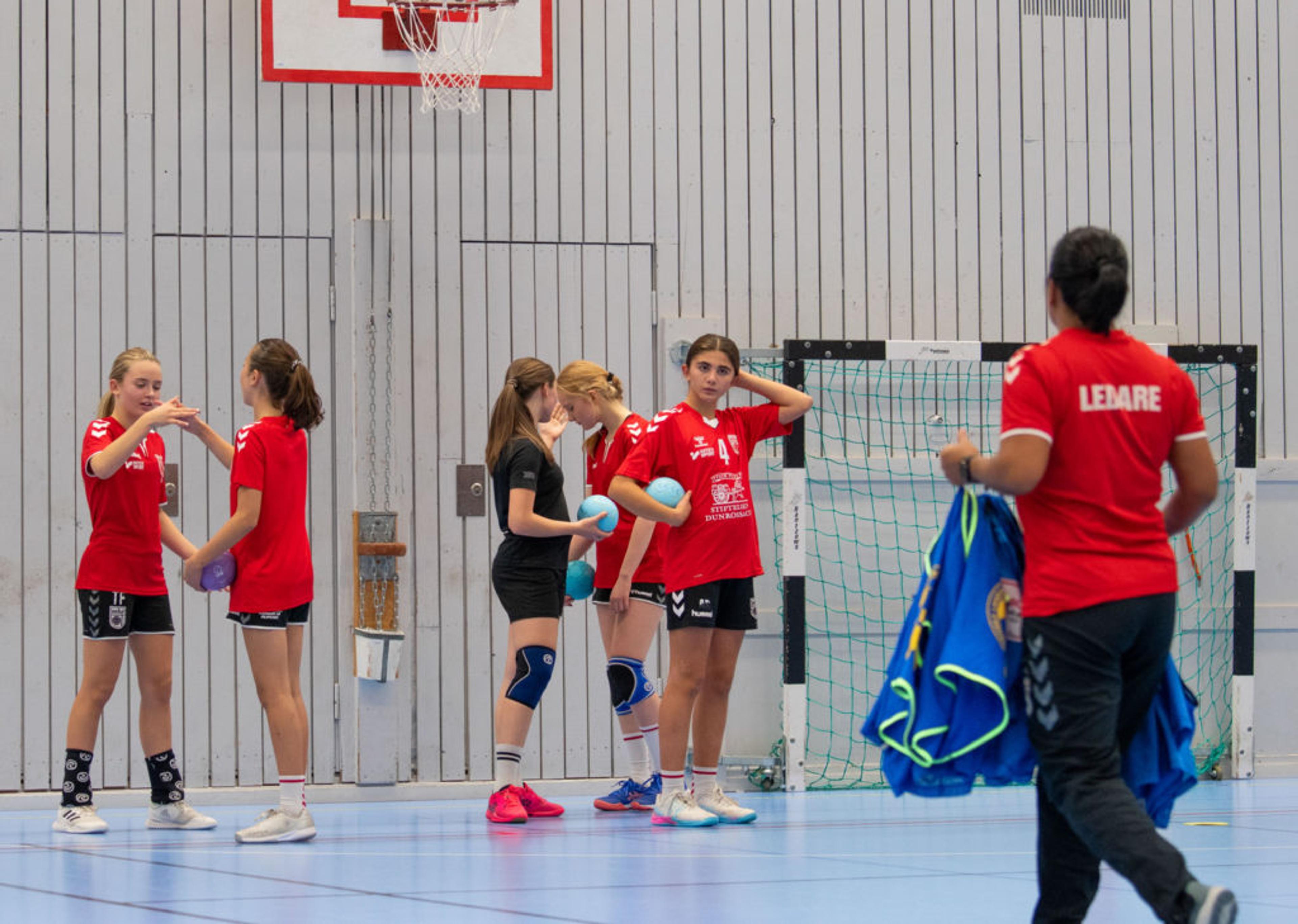 En grupp ungdomar i sportkläder samlas i en gymnastikhall med en basketkorg och innebandymål i bakgrunden. En ledare bär en röd tröja där det står "LEDARE" och håller i blå västar.