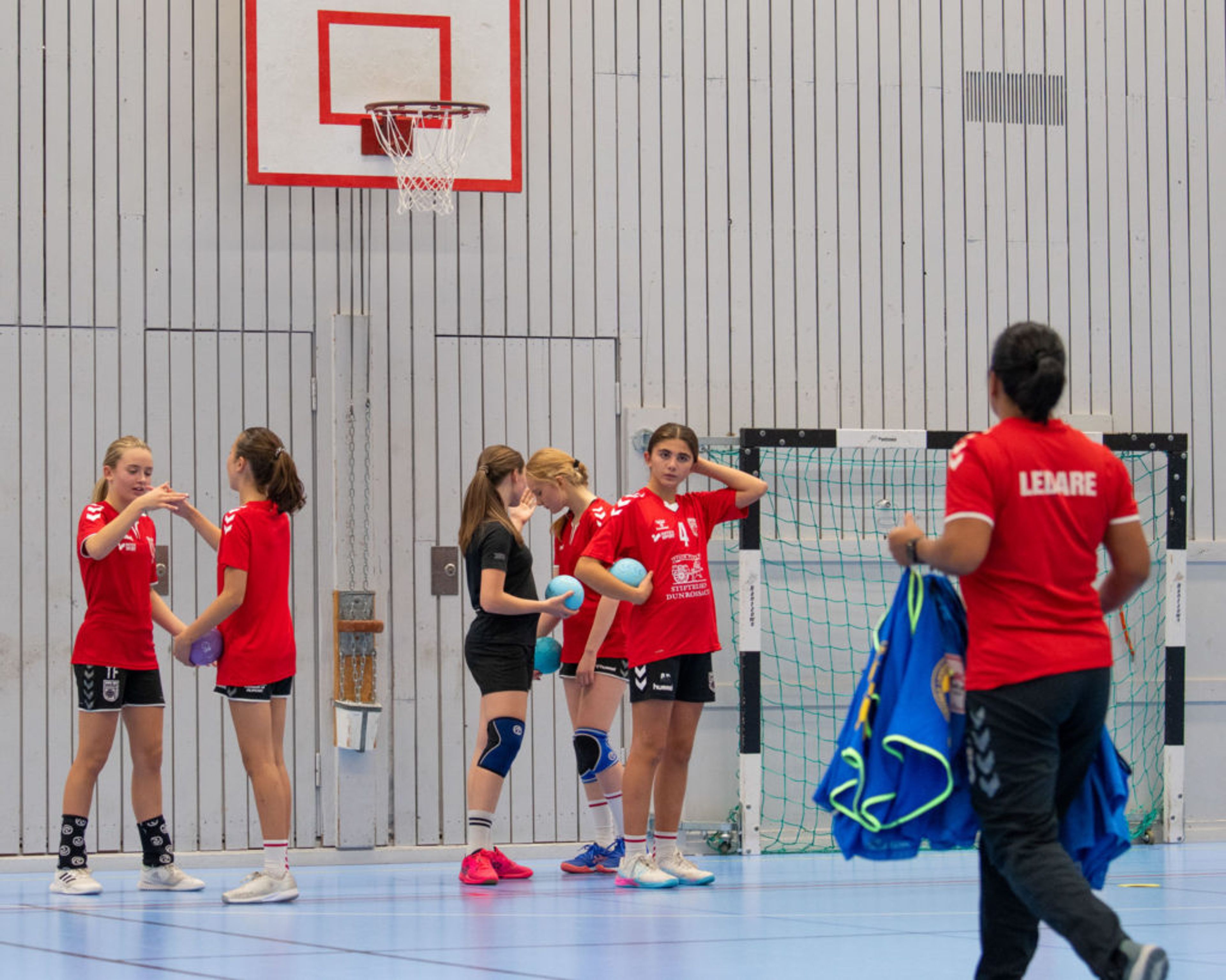 En grupp ungdomar i sportkläder samlas i en gymnastikhall med en basketkorg och innebandymål i bakgrunden. En ledare bär en röd tröja där det står "LEDARE" och håller i blå västar.