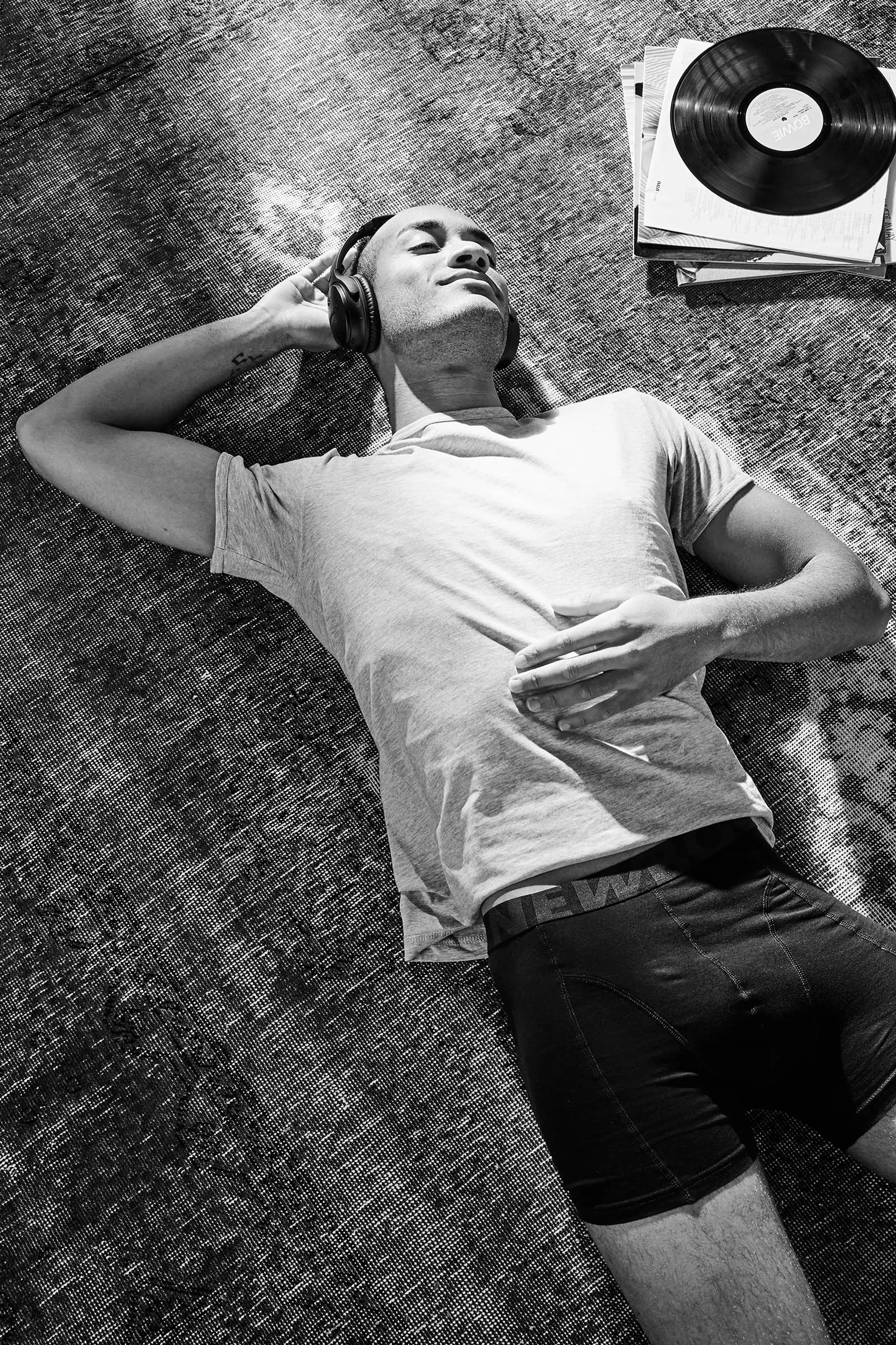 En person ligger avslappnat på en matta med hörlurar på och en t-shirt tillsammans med svarta, åtsittande boxer-kalsonger. Intill ligger en hög vinylskivor med en skiva överst.