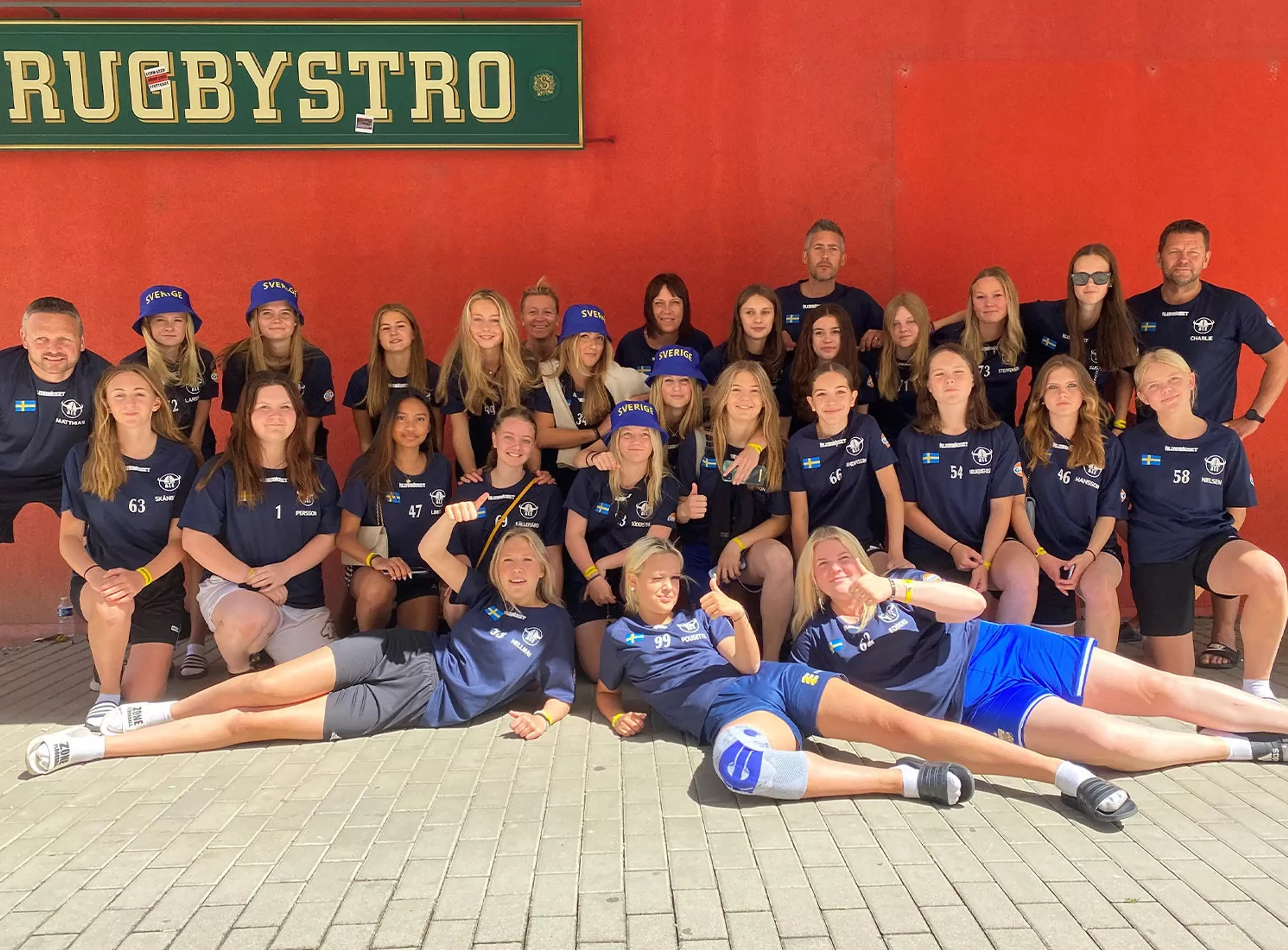 En stor grupp ungdomar och vuxna i matchkläder poserar tillsammans framför en röd vägg med en skylt där det står "Rugbystro". Några av ungdomarna bär blå hattar med texten "Sverige".