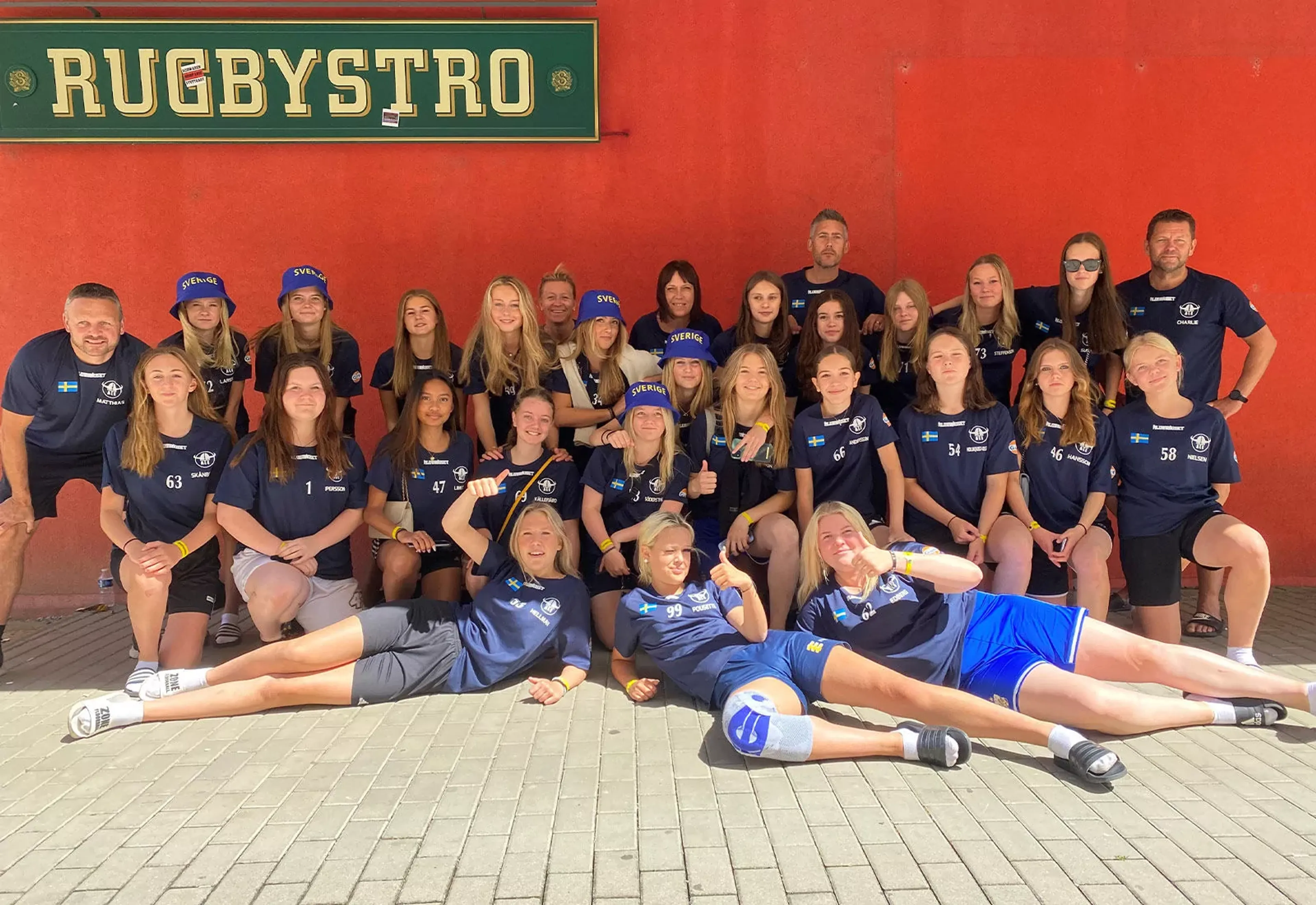 En stor grupp ungdomar och vuxna i matchkläder poserar tillsammans framför en röd vägg med en skylt där det står "Rugbystro". Några av ungdomarna bär blå hattar med texten "Sverige".