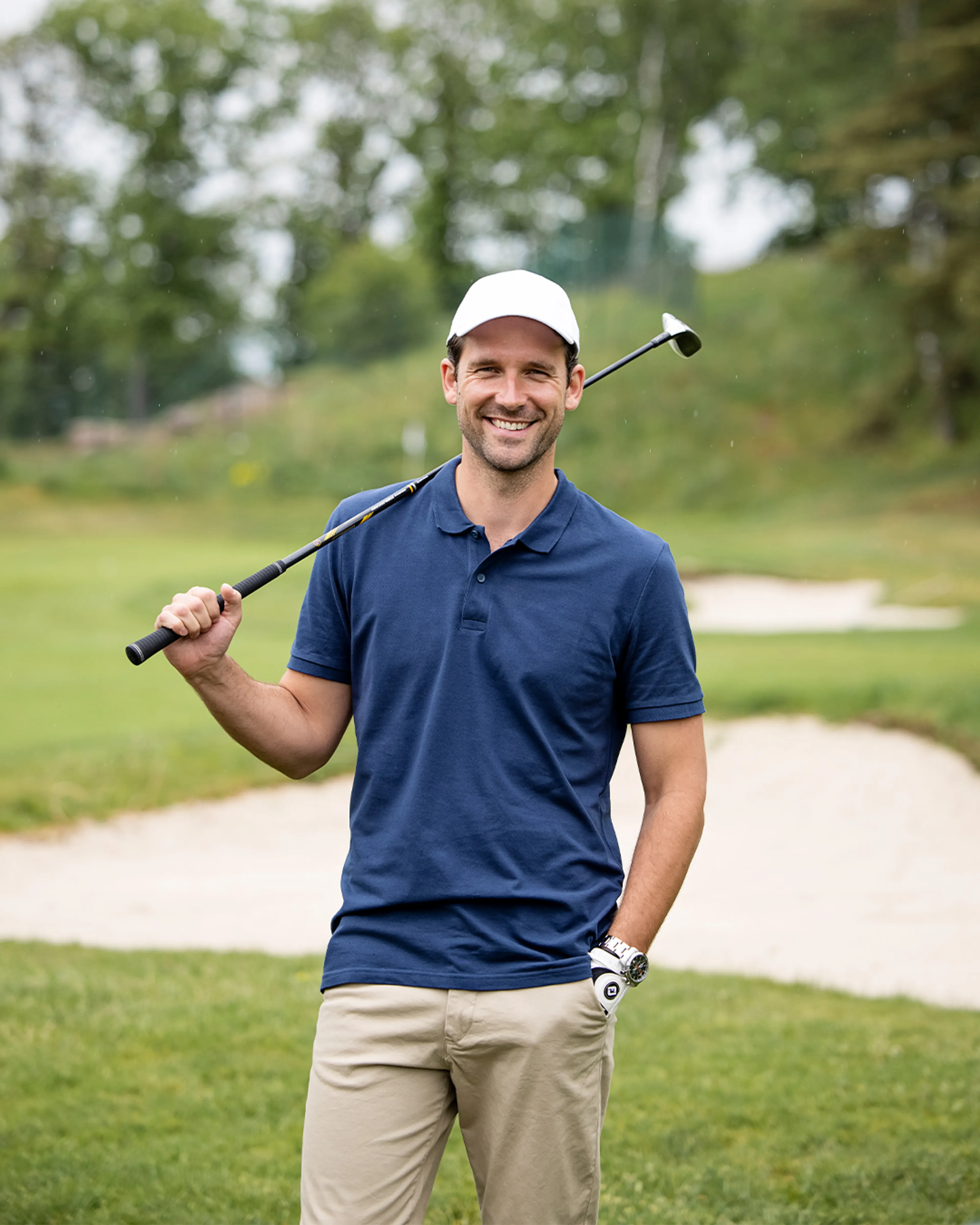 En man i beige byxor och mörkblå pikétröja håller en golfklubba över axeln på en golfbana. Han bär vit keps och har ett leende på läpparna.