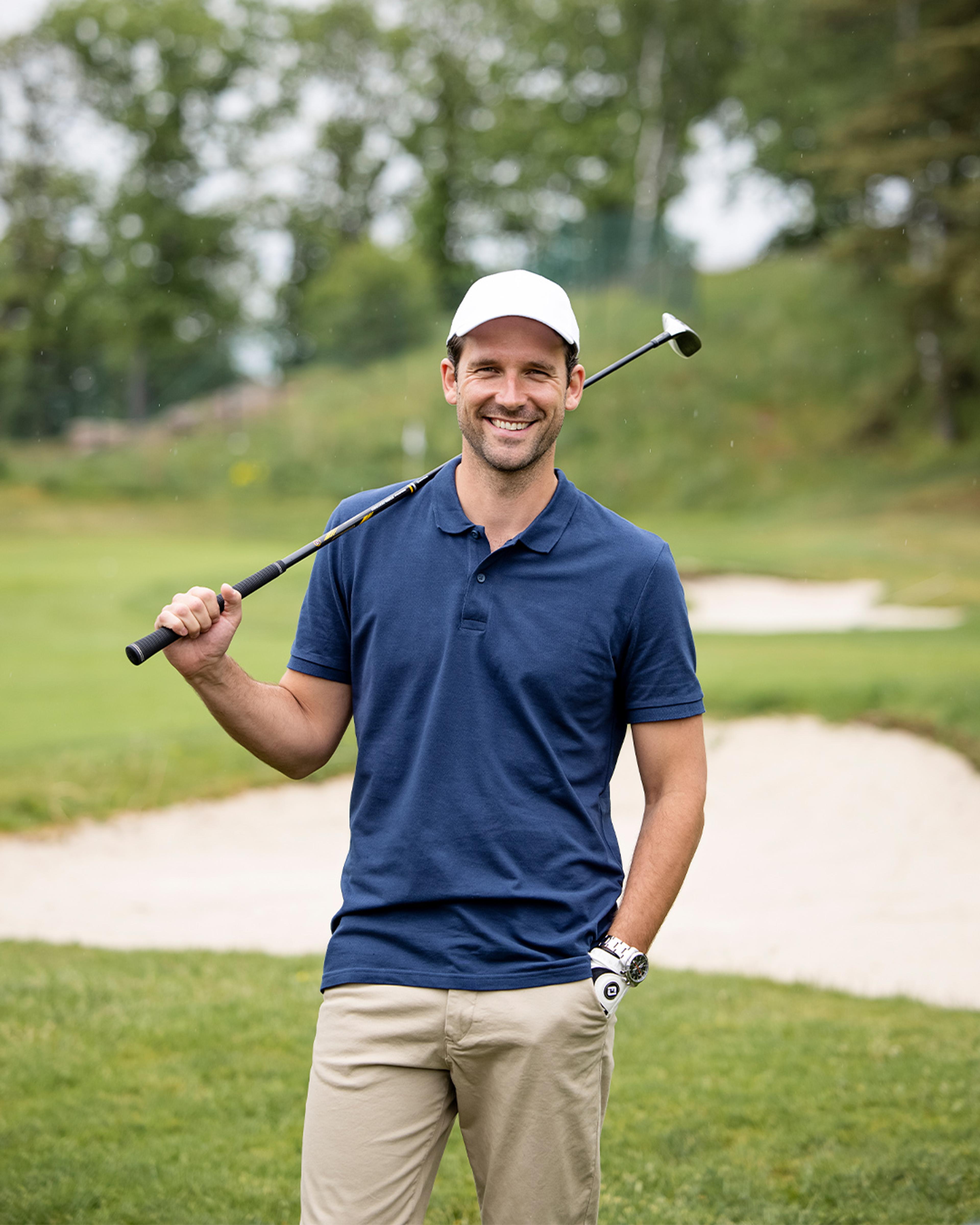 En man i beige byxor och mörkblå pikétröja håller en golfklubba över axeln på en golfbana. Han bär vit keps och har ett leende på läpparna.