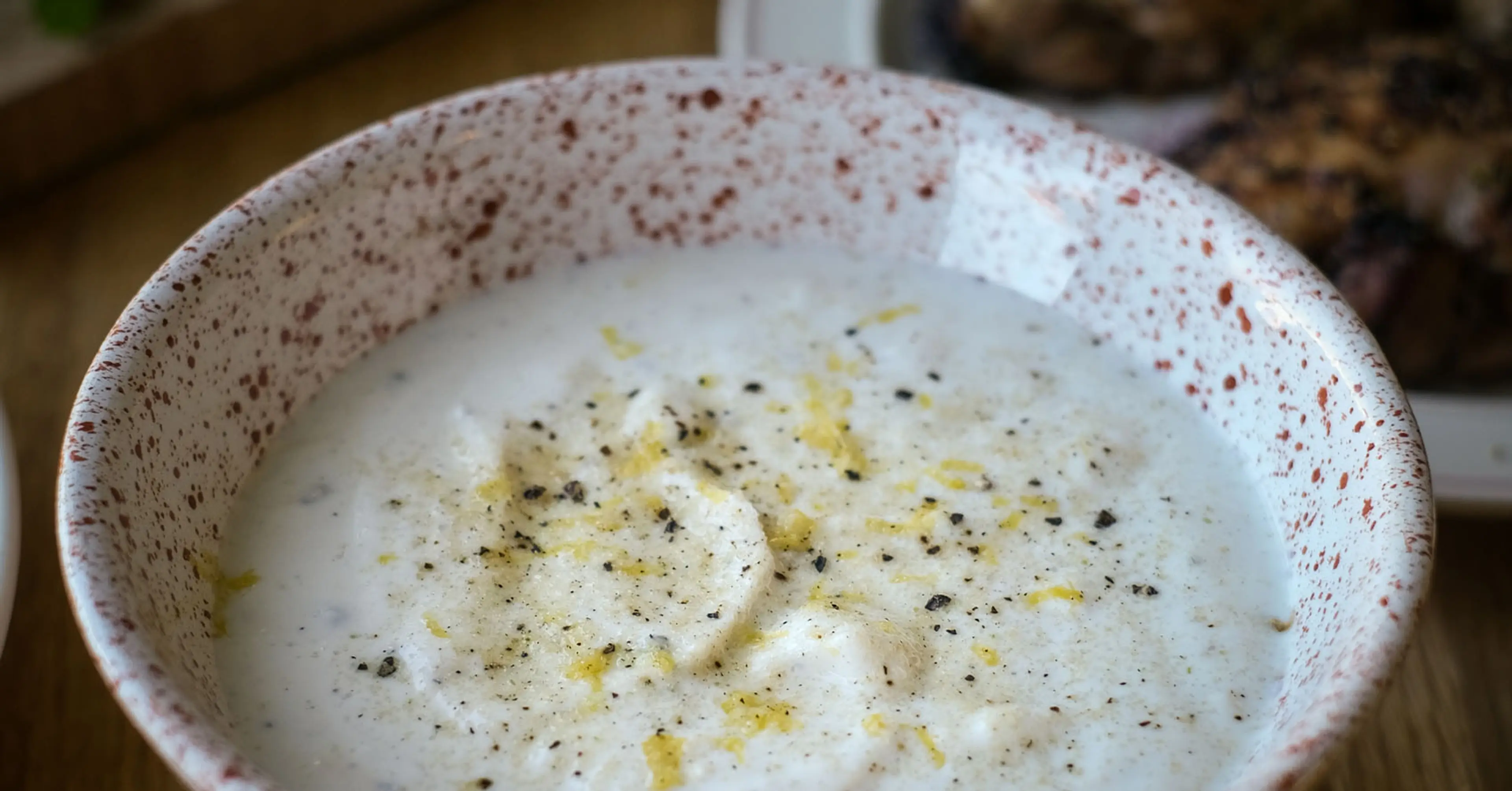 En skål med en krämig vit sås toppad med svartpeppar och citronskal. I bakgrunden syns limeklyftor och grillade bitar på en tallrik.