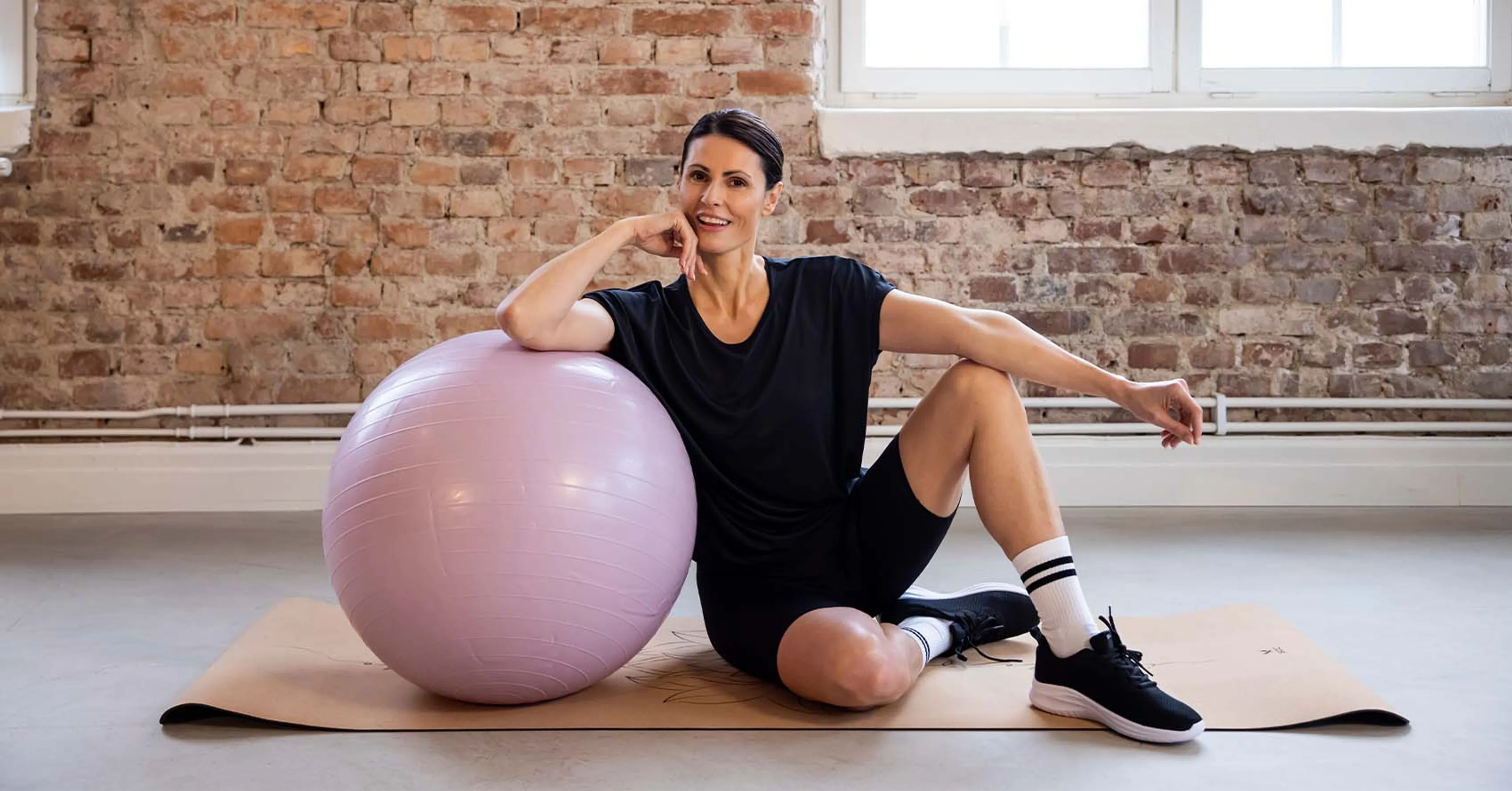 En kvinna i sportig svart outfit och svarta träningsskor sitter avslappnat på en yogamatta med en ljuslila pilatesboll vid sidan av sig. Bakgrunden är en vägg av röd tegelsten och ett stort fönster släpper in dags