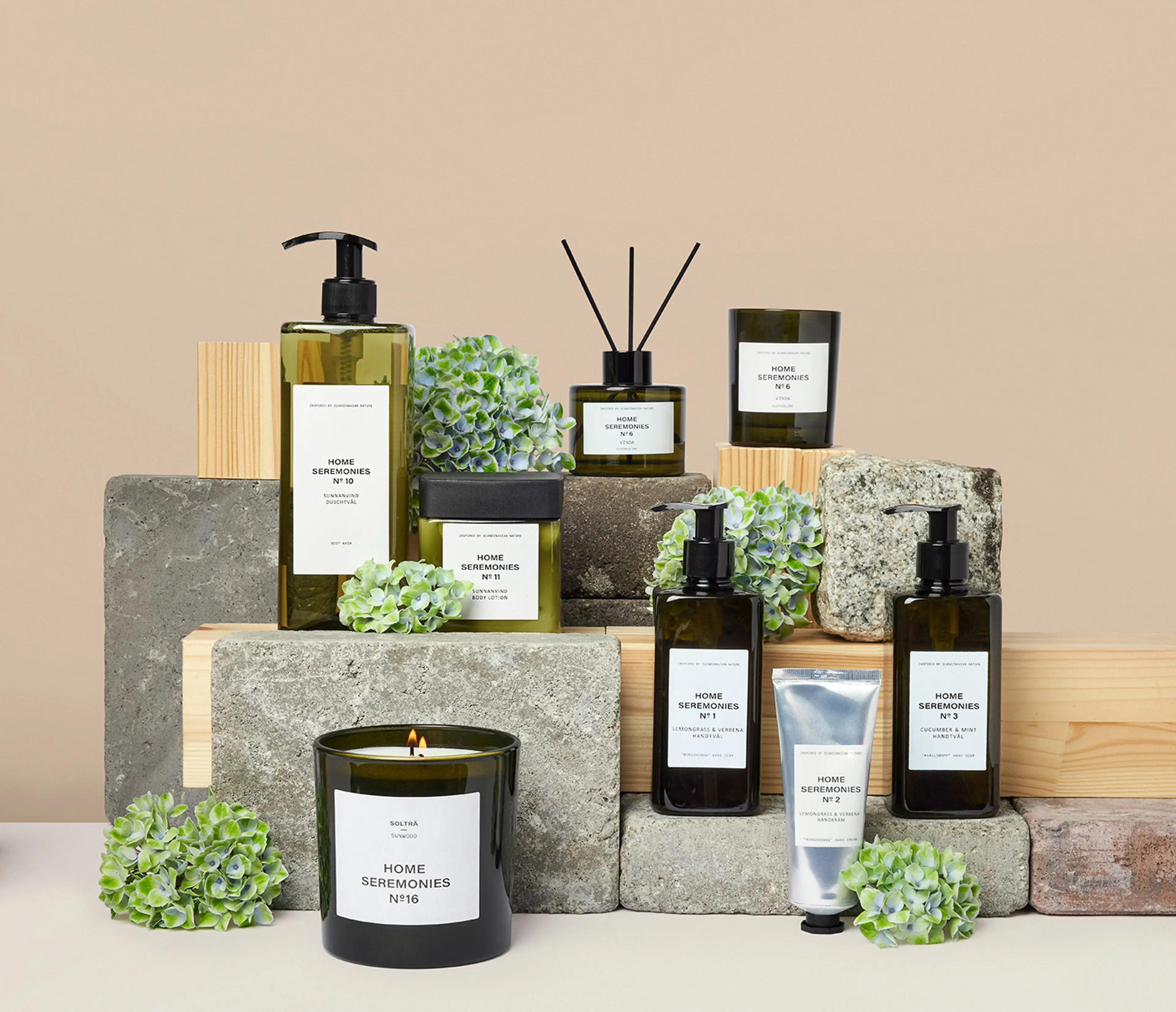 En samling doftljus, handtvålar, handkräm och body lotion i gröna glasflaskor med minimalistiska etiketter, arrangerade på trä- och stenkuber tillsammans med gröna blomdekorationer. Produkten ger en känsla av fräschhet