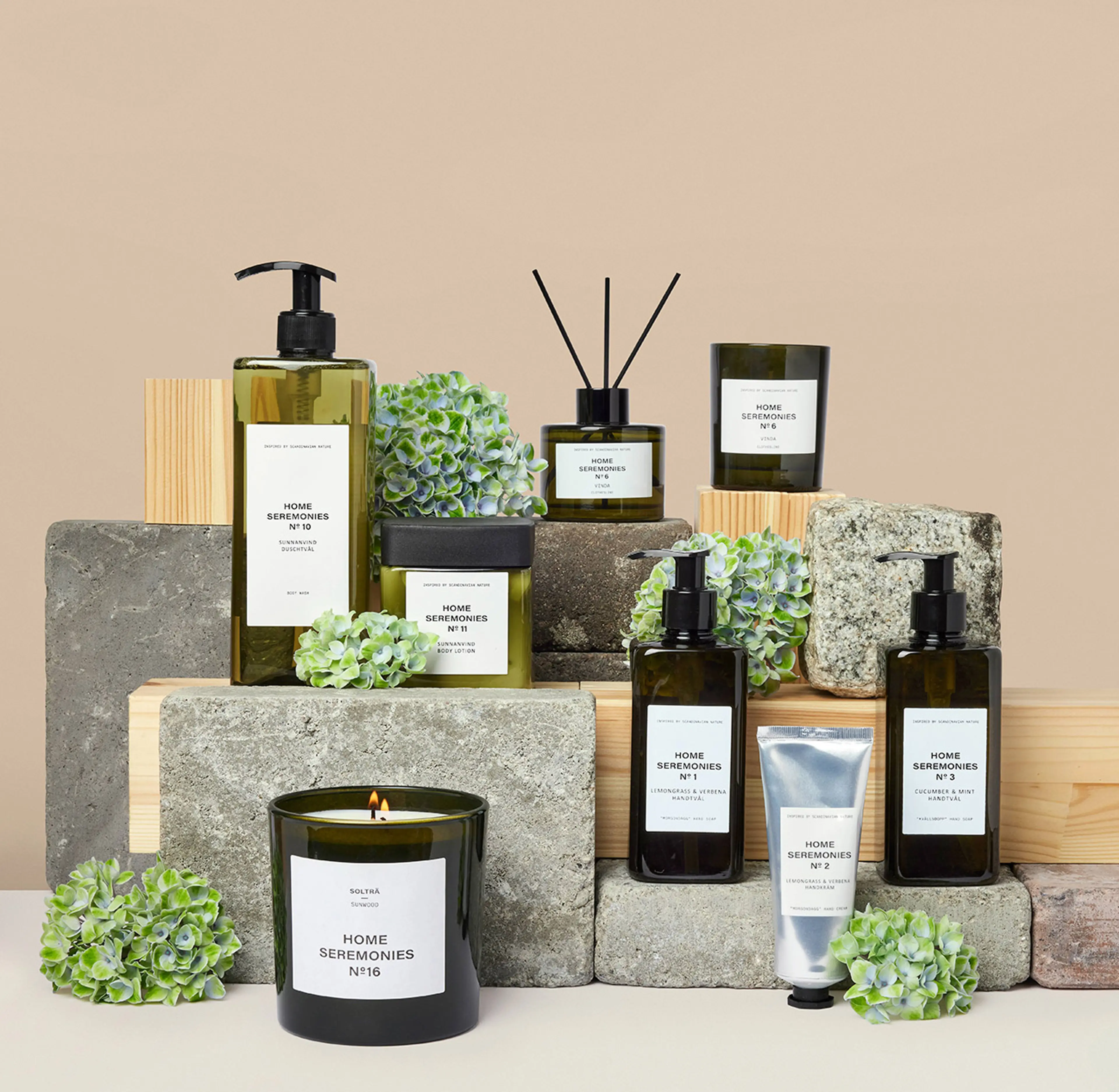 En samling doftljus, handtvålar, handkräm och body lotion i gröna glasflaskor med minimalistiska etiketter, arrangerade på trä- och stenkuber tillsammans med gröna blomdekorationer. Produkten ger en känsla av fräschhet