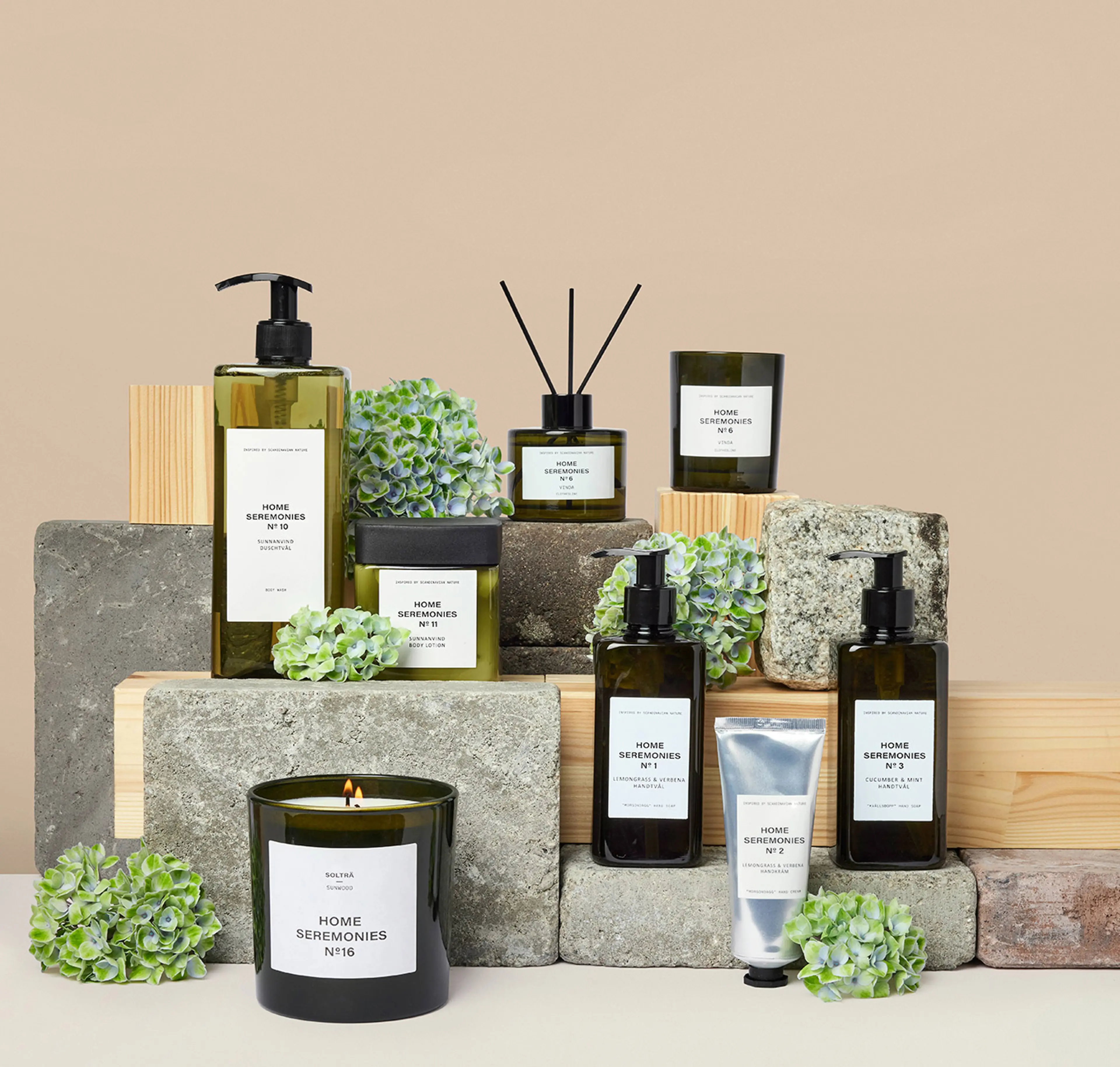 En samling doftljus, handtvålar, handkräm och body lotion i gröna glasflaskor med minimalistiska etiketter, arrangerade på trä- och stenkuber tillsammans med gröna blomdekorationer. Produkten ger en känsla av fräschhet
