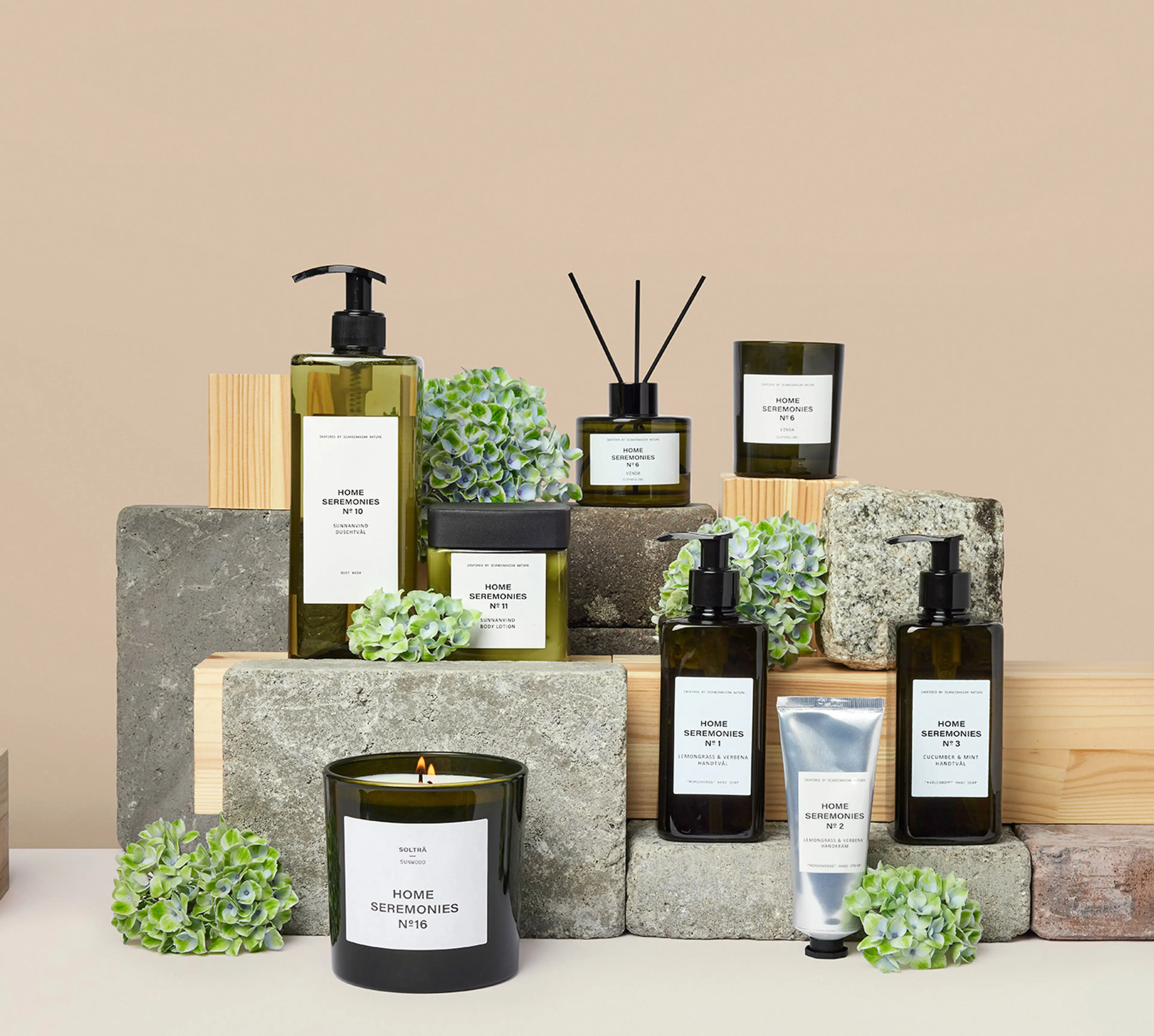 En samling doftljus, handtvålar, handkräm och body lotion i gröna glasflaskor med minimalistiska etiketter, arrangerade på trä- och stenkuber tillsammans med gröna blomdekorationer. Produkten ger en känsla av fräschhet