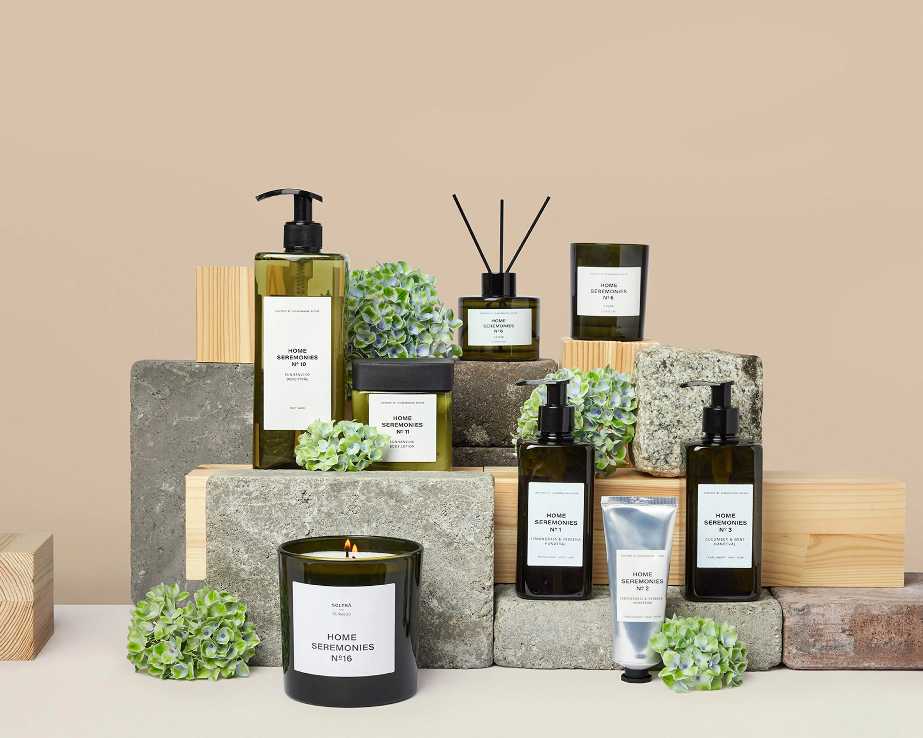 En samling doftljus, handtvålar, handkräm och body lotion i gröna glasflaskor med minimalistiska etiketter, arrangerade på trä- och stenkuber tillsammans med gröna blomdekorationer. Produkten ger en känsla av fräschhet