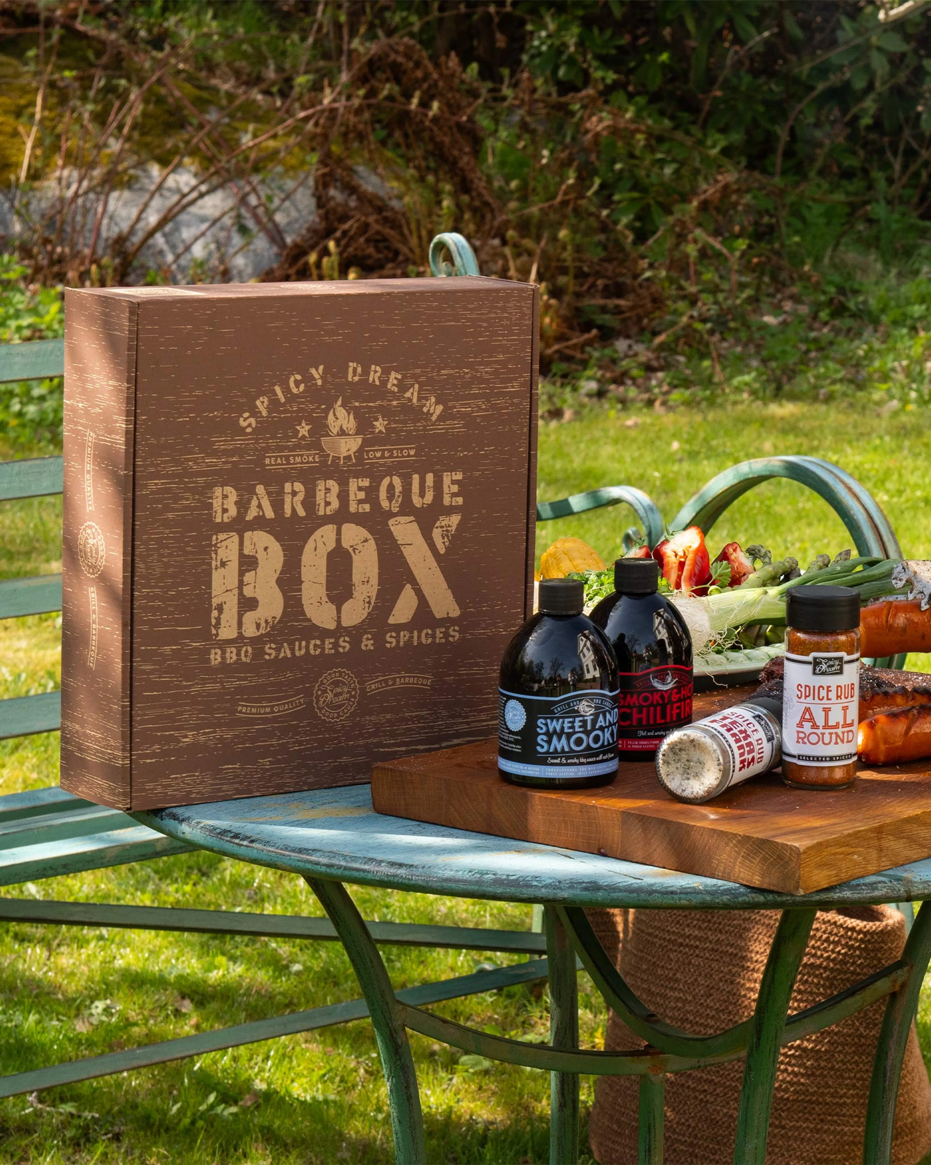 En brun presentförpackning märkt "Barbeque Box" står på ett blågrönt metallbord bredvid en skärbräda med två flaskor sås och två kryddburkar. I bakgrunden syns färska grönsaker och gräsmatta