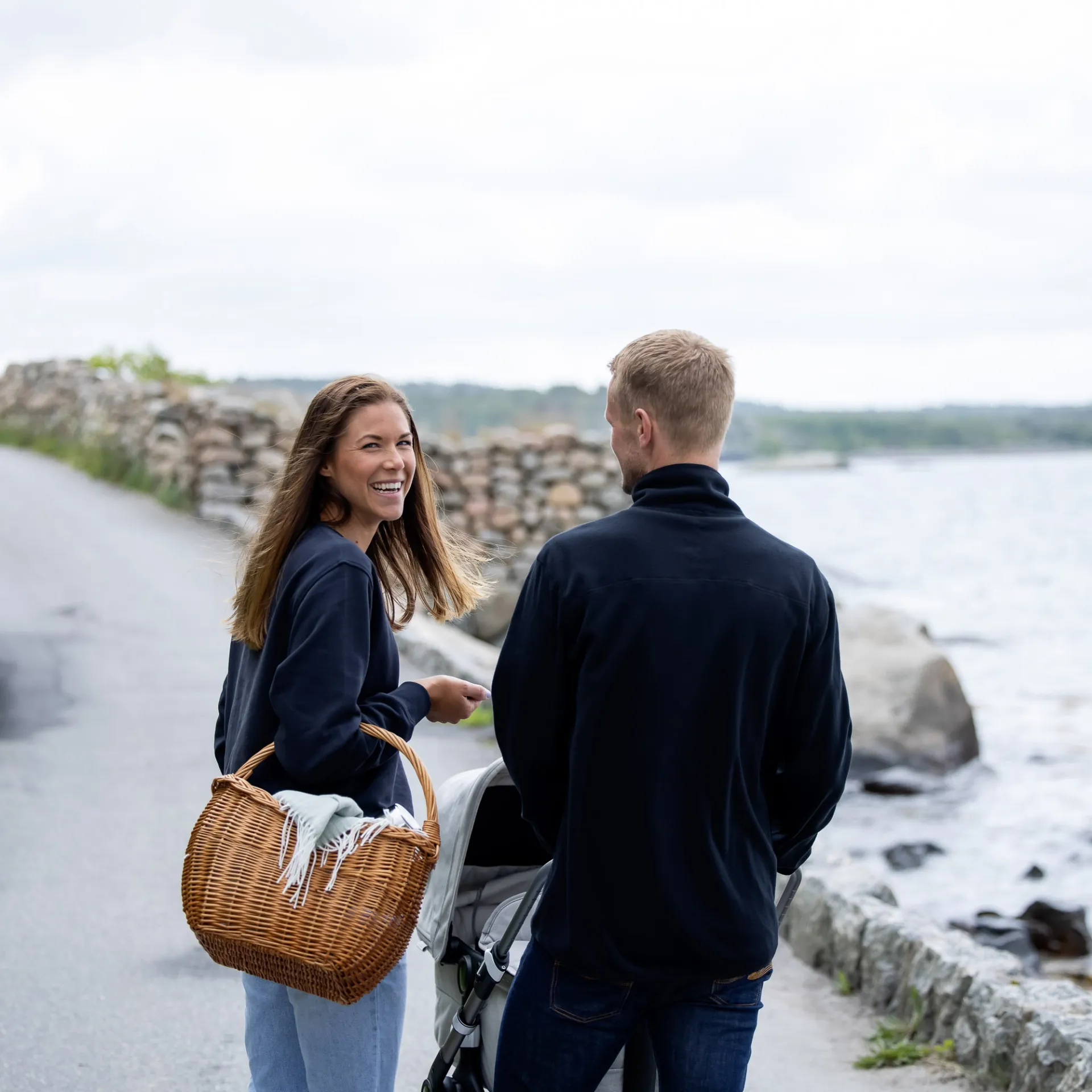 En kvinna i marinblå sweatshirt med ljusa jeans håller en flätad korg, och en man i mörk fleecejacka går bredvid henne med en barnvagn vid en stenig kustväg.