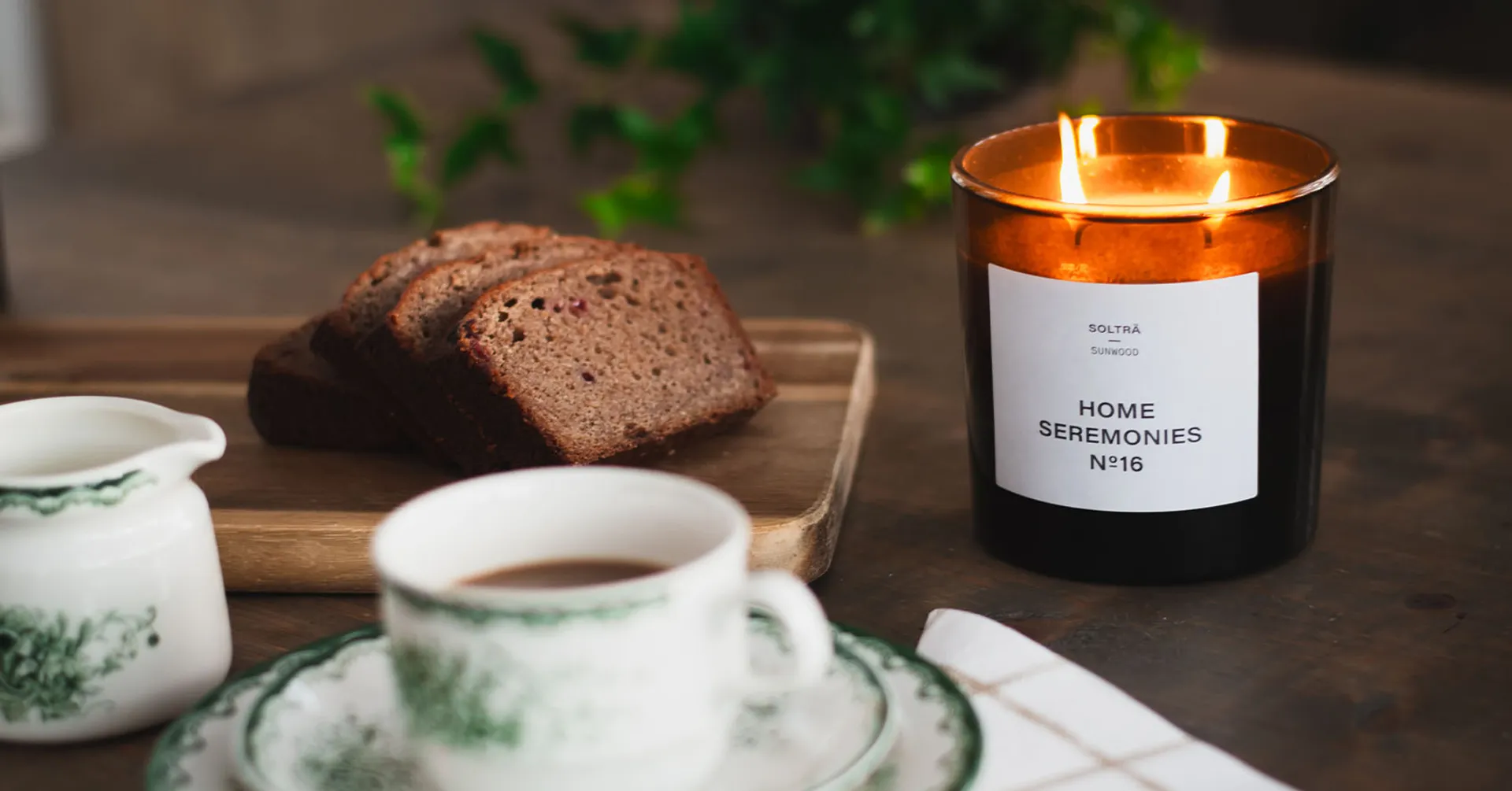 Doftljus från Home Seremonies på ett dukat mörk träbord med en kaffekopp och några bitar mjuk pepparkaka