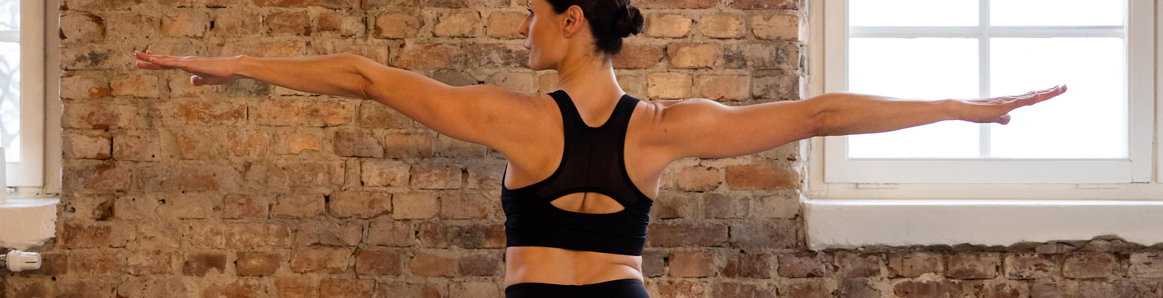 En svart sport-bh med breda axelband och ett cut out-detalj på ryggen, perfekt för högintensiv träning. Modellen står i en träningsposition med armarna utsträckta i ett rustikt rum med tegelvägg och stora fön