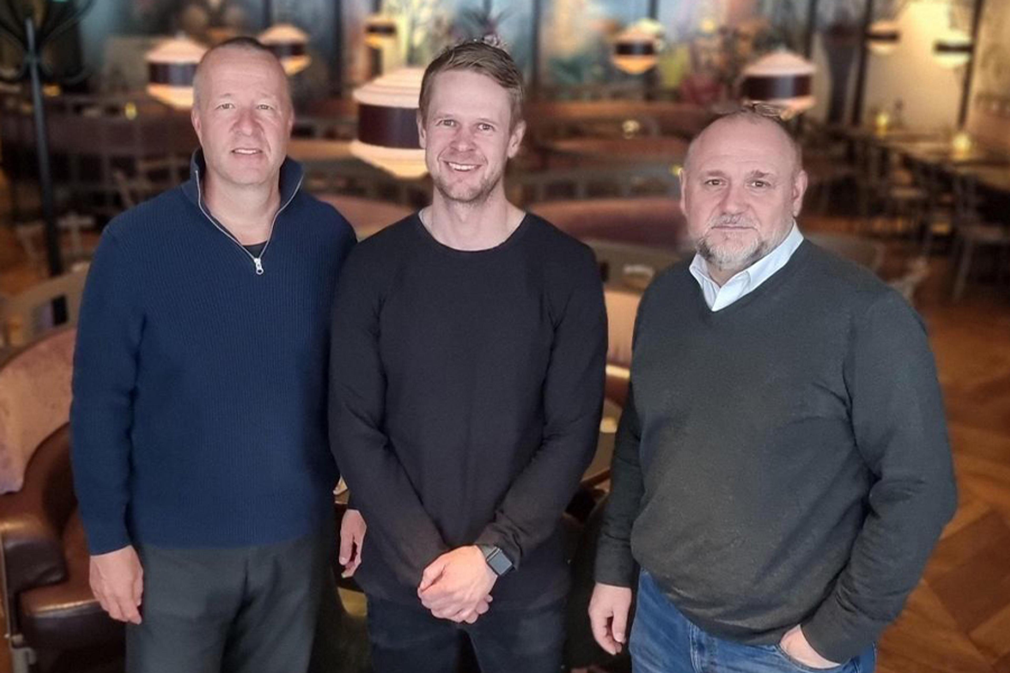 Tre män i tröjor står tillsammans i en mysig och varm inomhusmiljö med mjuk belysning och trämöbler runt omkring.