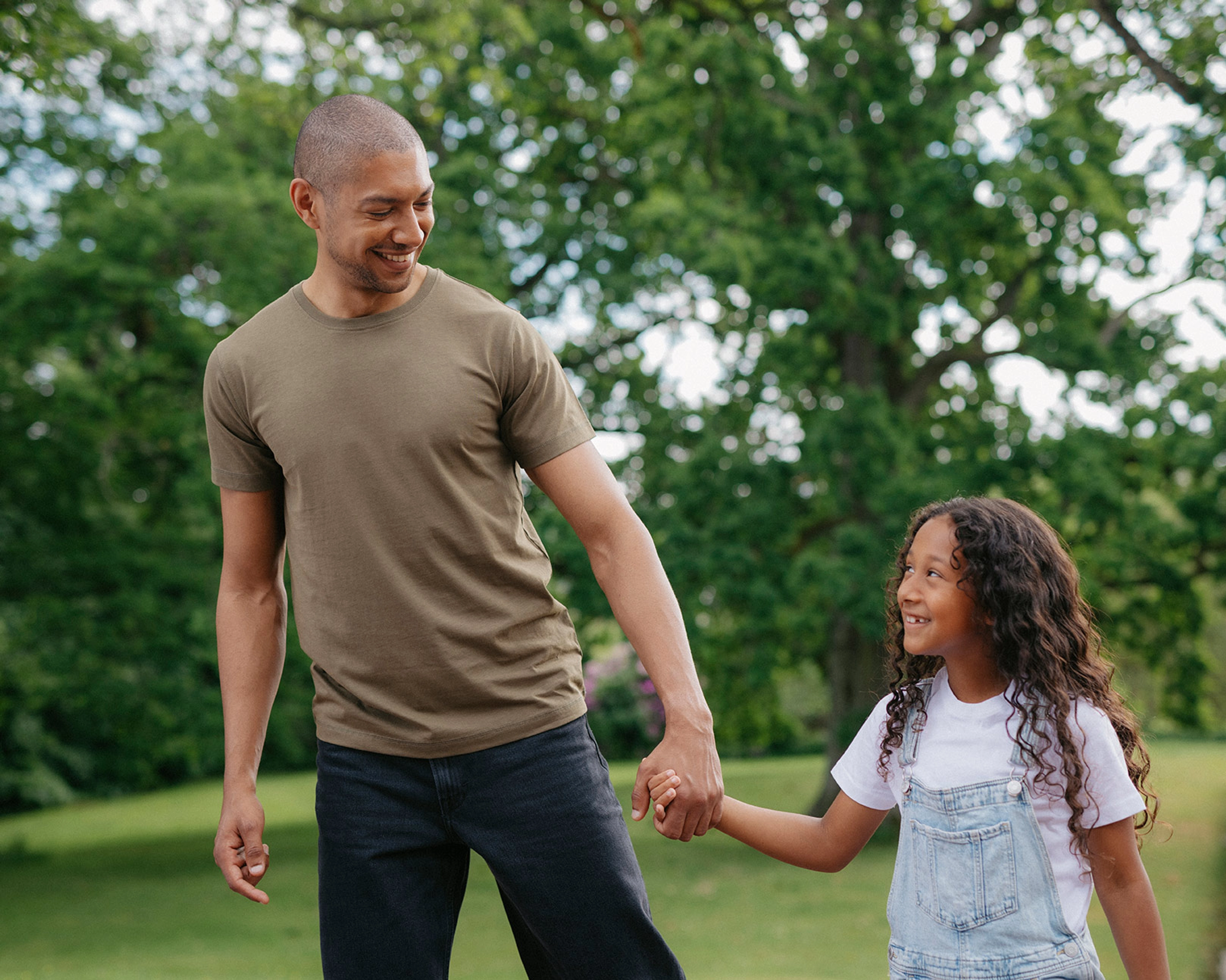 En man i en enkel, olivgrön t-shirt håller handen med ett leende barn i vit t-shirt och ljusa hängselbyxor utomhus bland gröna träd.