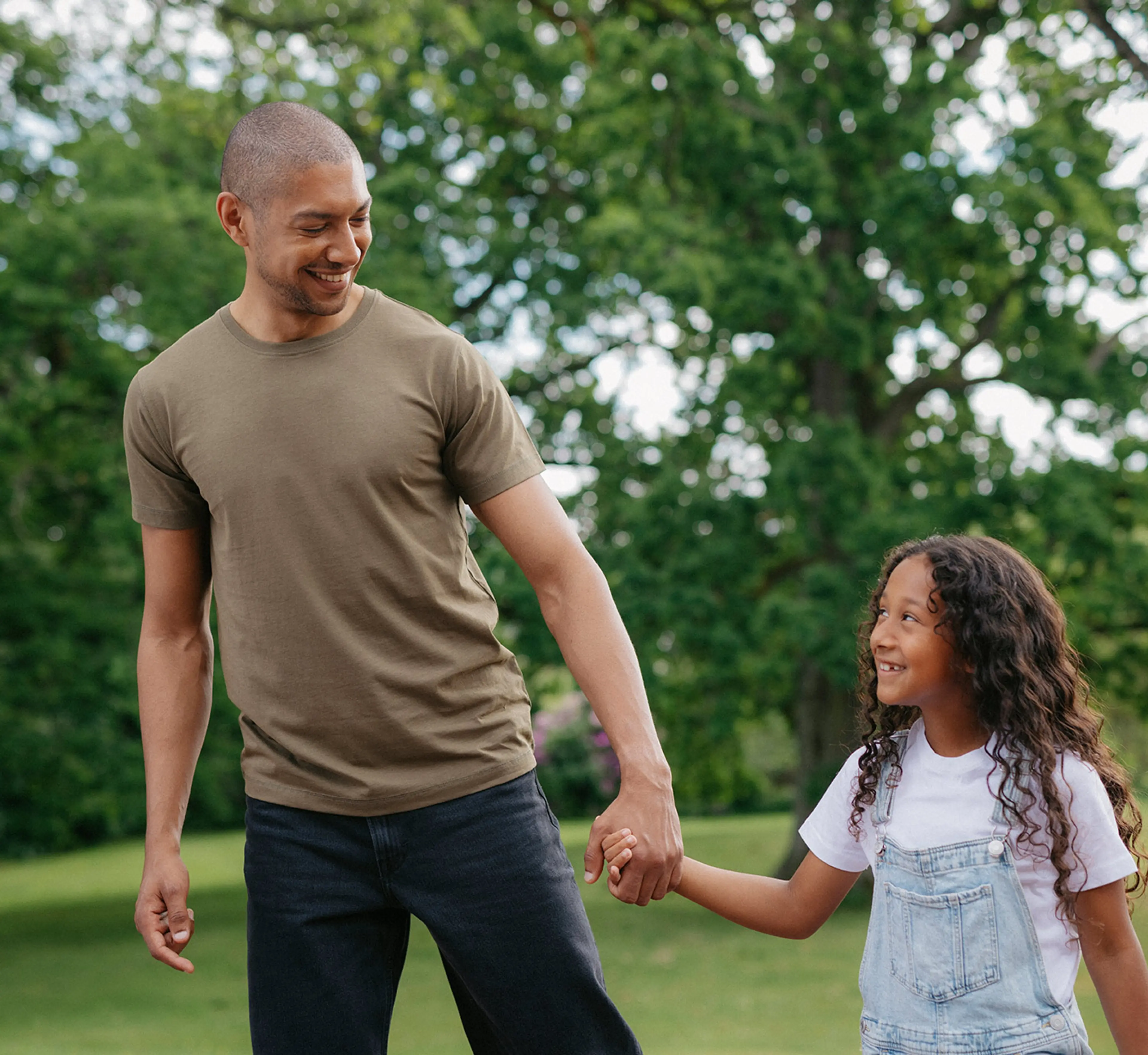 En man i en enkel, olivgrön t-shirt håller handen med ett leende barn i vit t-shirt och ljusa hängselbyxor utomhus bland gröna träd.