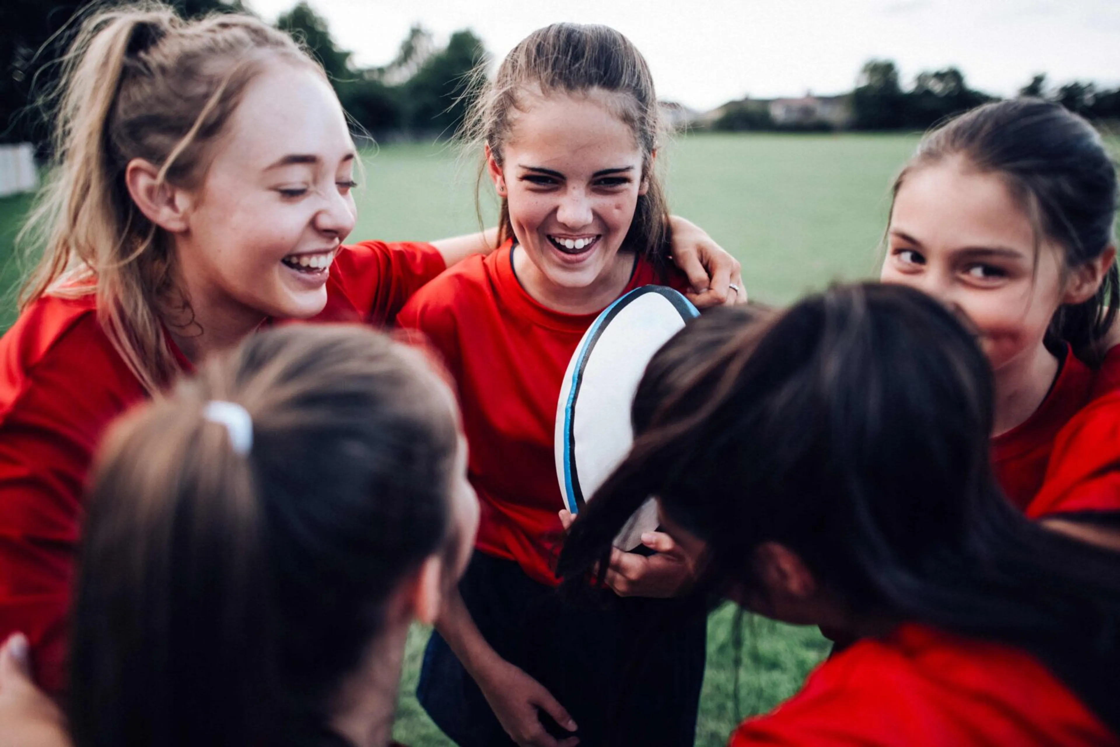 Fem tjejer i röda tröjor samlas i en grupp på en gräsplan, där en av dem håller i en rugbyboll. De ser glada och sammansvetsade ut.