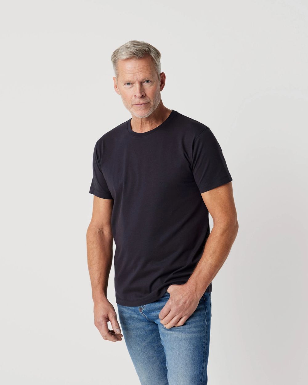 En enkel, mörkblå t-shirt med rund hals och korta ärmar, bärs av en person med ljus hy och grått hår. Tröjan har en rak och avslappnad passform.