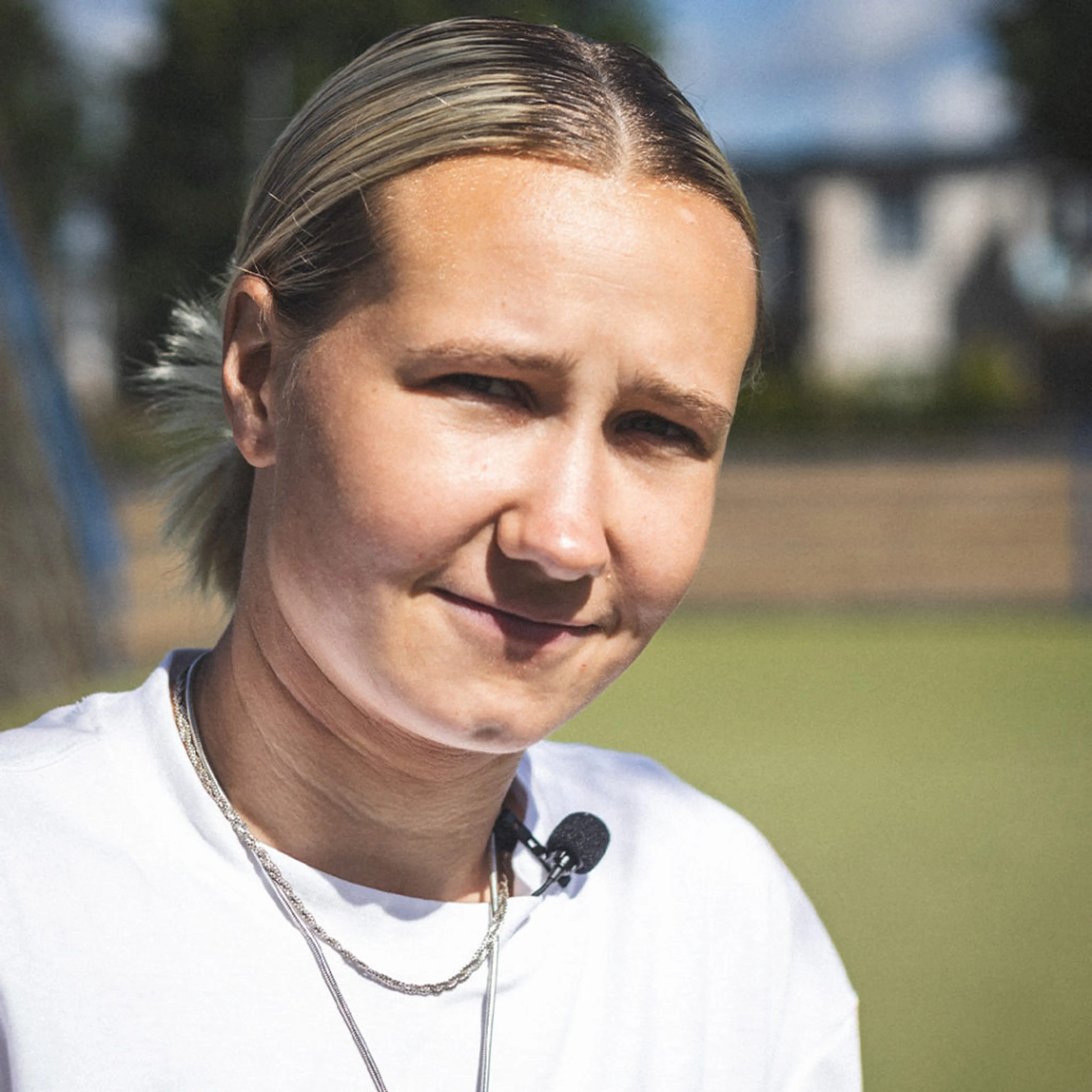 En person med ljust uppsatt hår och vit t-shirt bär en liten mikrofonfästen vid halsen, fotograferad utomhus i dagsljus. Bakgrunden är suddig med inslag av grönska och byggnader.
