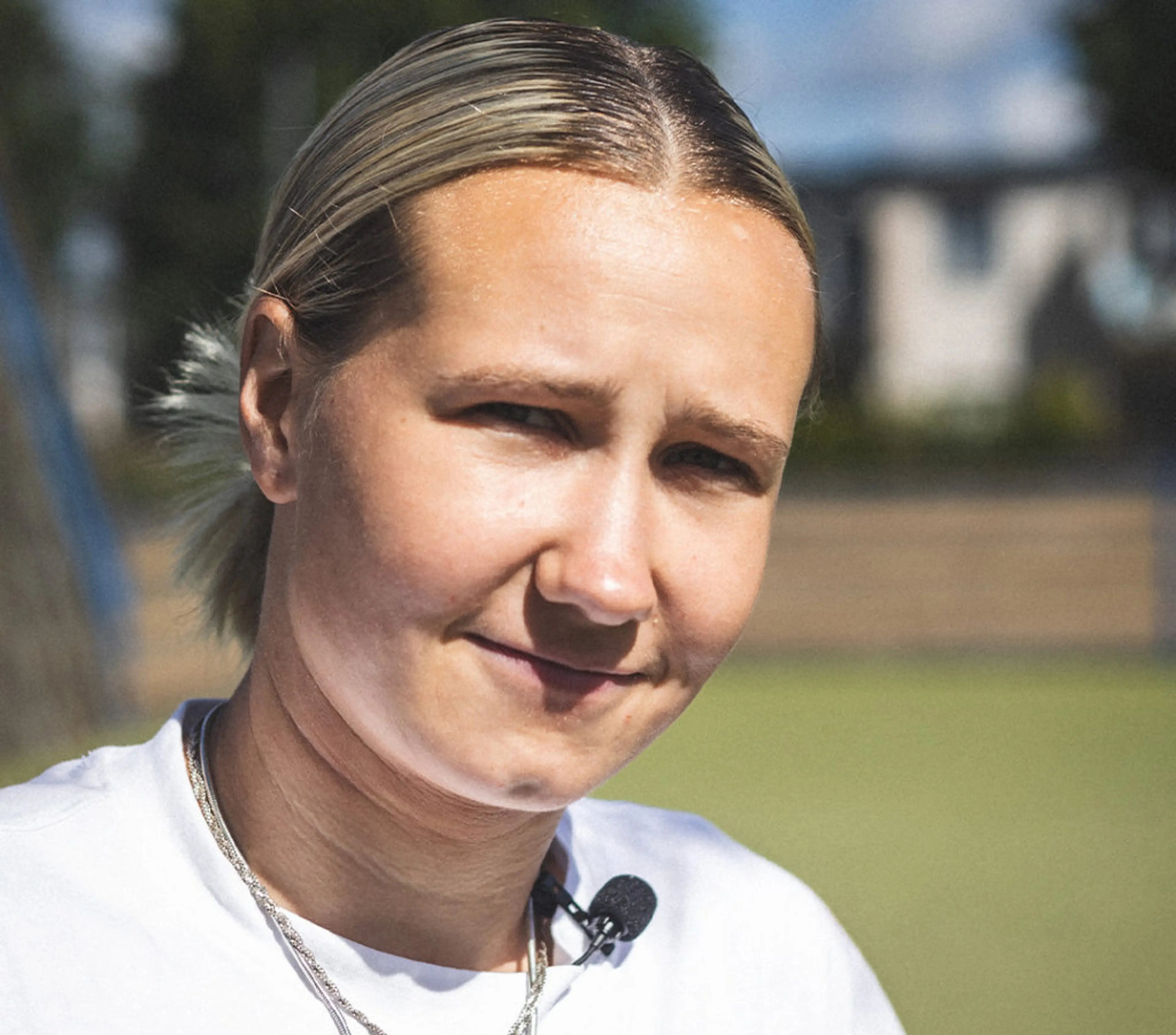 En person med ljust uppsatt hår och vit t-shirt bär en liten mikrofonfästen vid halsen, fotograferad utomhus i dagsljus. Bakgrunden är suddig med inslag av grönska och byggnader.
