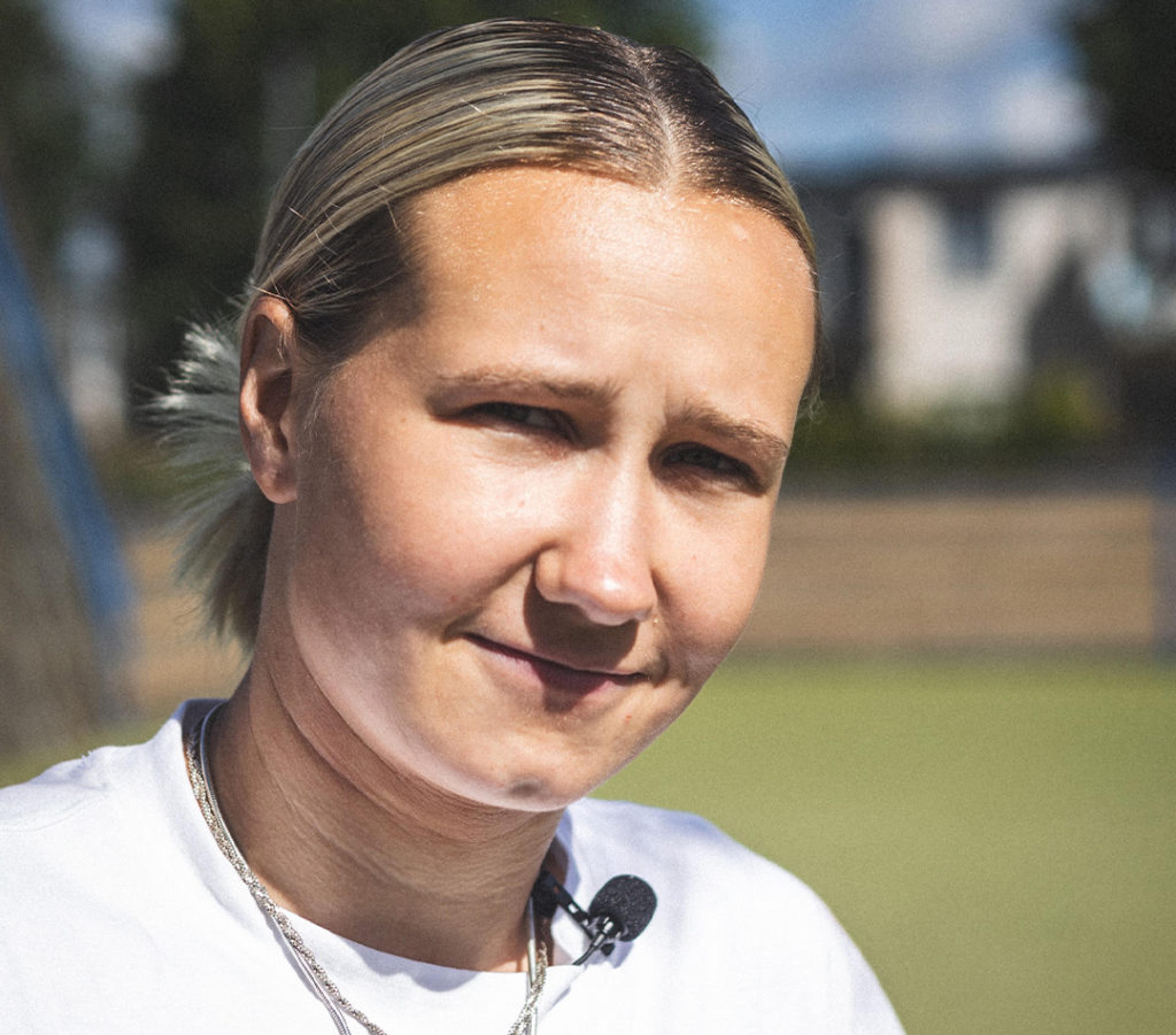 En person med ljust uppsatt hår och vit t-shirt bär en liten mikrofonfästen vid halsen, fotograferad utomhus i dagsljus. Bakgrunden är suddig med inslag av grönska och byggnader.