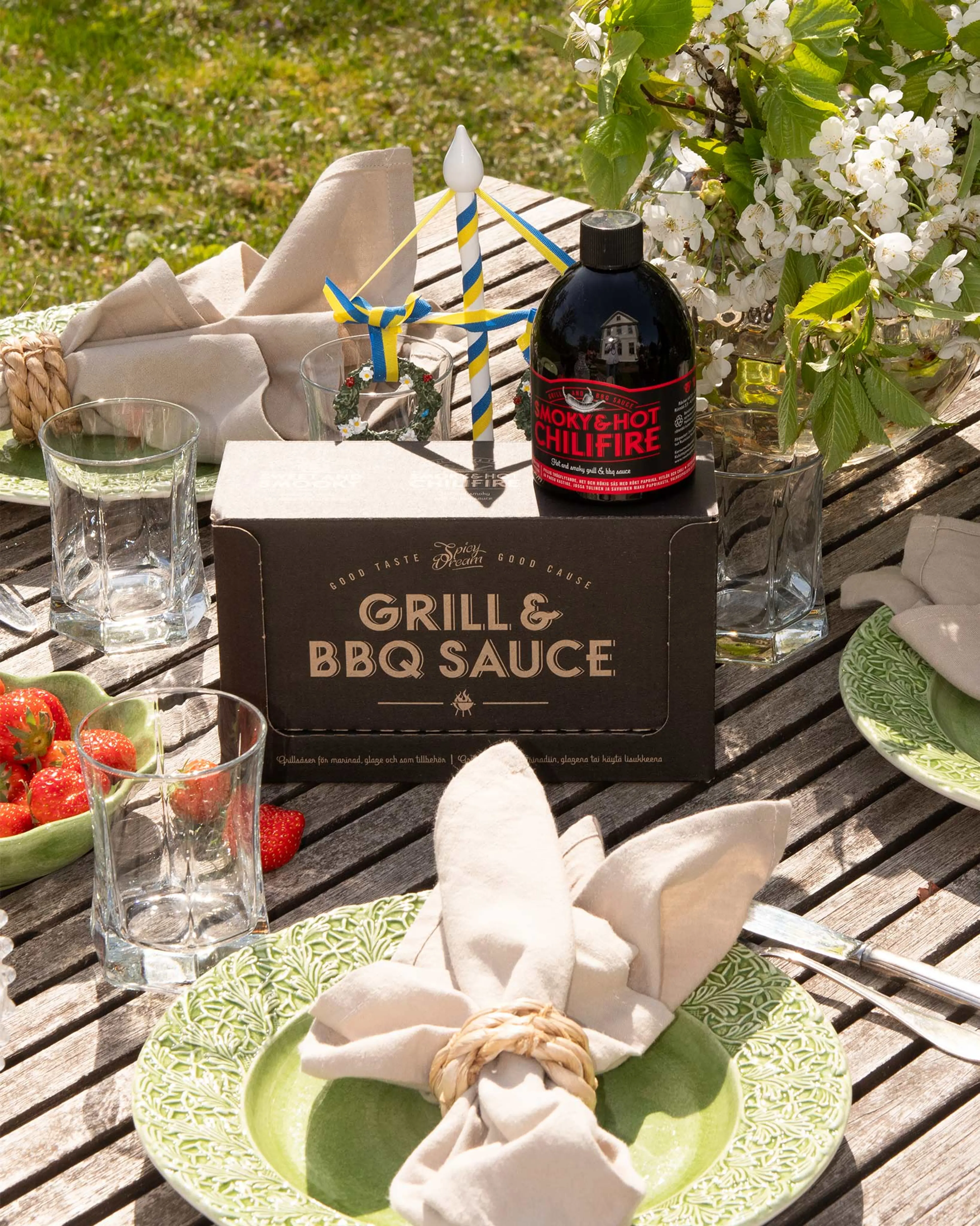 En flaska kryddig BBQ-sås står på en svart förpackning med texten "Grill & BBQ Sauce" på ett träbord. Bordet är dukat med gröna tallrikar, linneservetter och ett litet blågult midsommarstångs