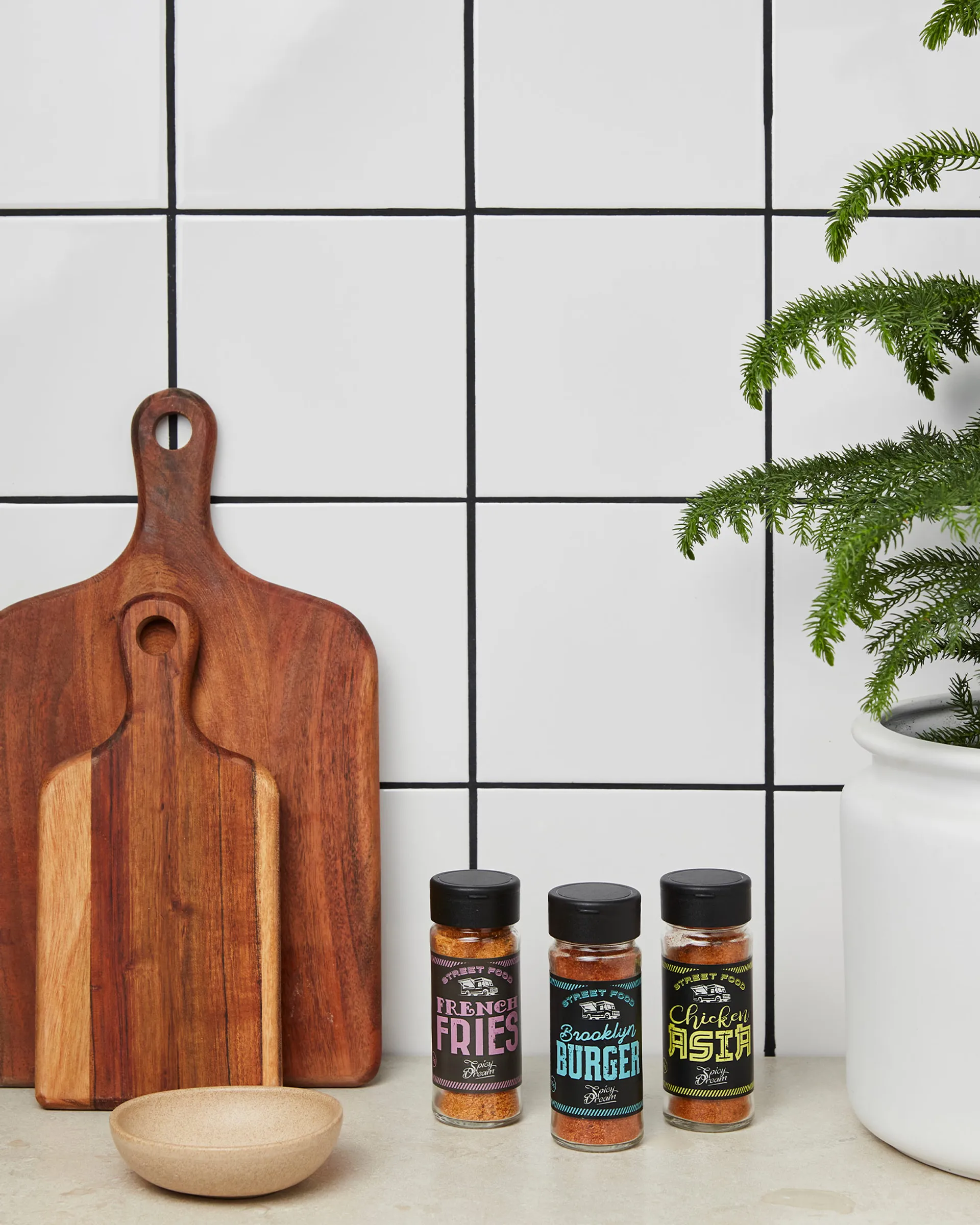 Tre kryddburkar med smakerna French Fries, Brooklyn Burger och Chicken Pasta står på en köksbänk bredvid två träskärbrädor och en grön växt i vit kruka.