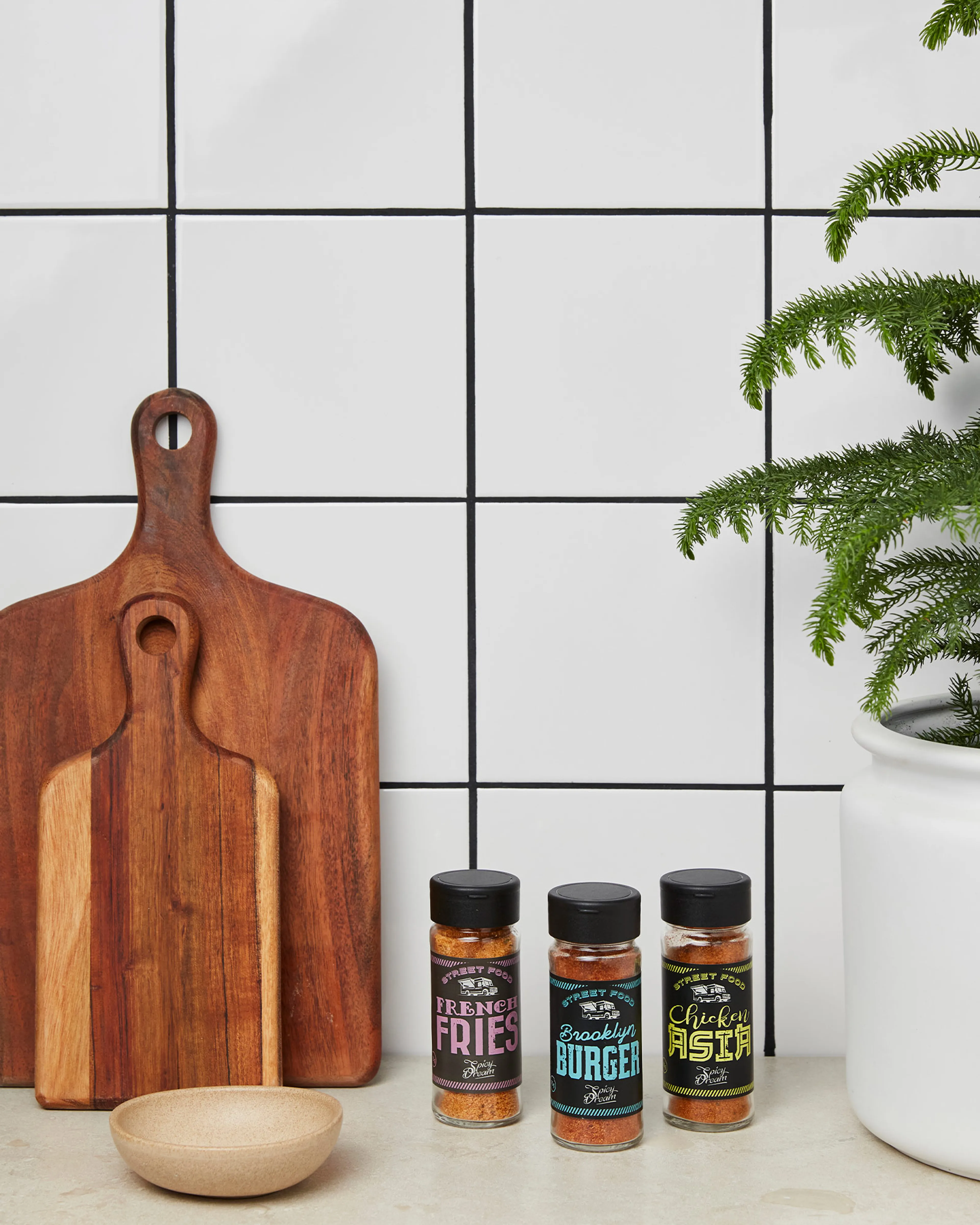 Tre kryddburkar med smakerna French Fries, Brooklyn Burger och Chicken Pasta står på en köksbänk bredvid två träskärbrädor och en grön växt i vit kruka.