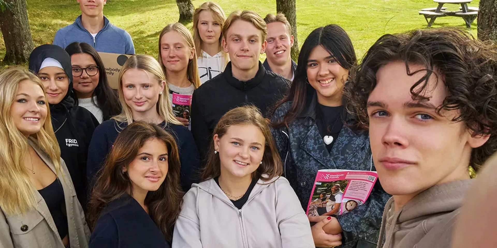 En grupp gymnasieelever samlade utomhus på gräsmatta, några håller broschyrer. De ler och ser avslappnade ut i en naturlig miljö.